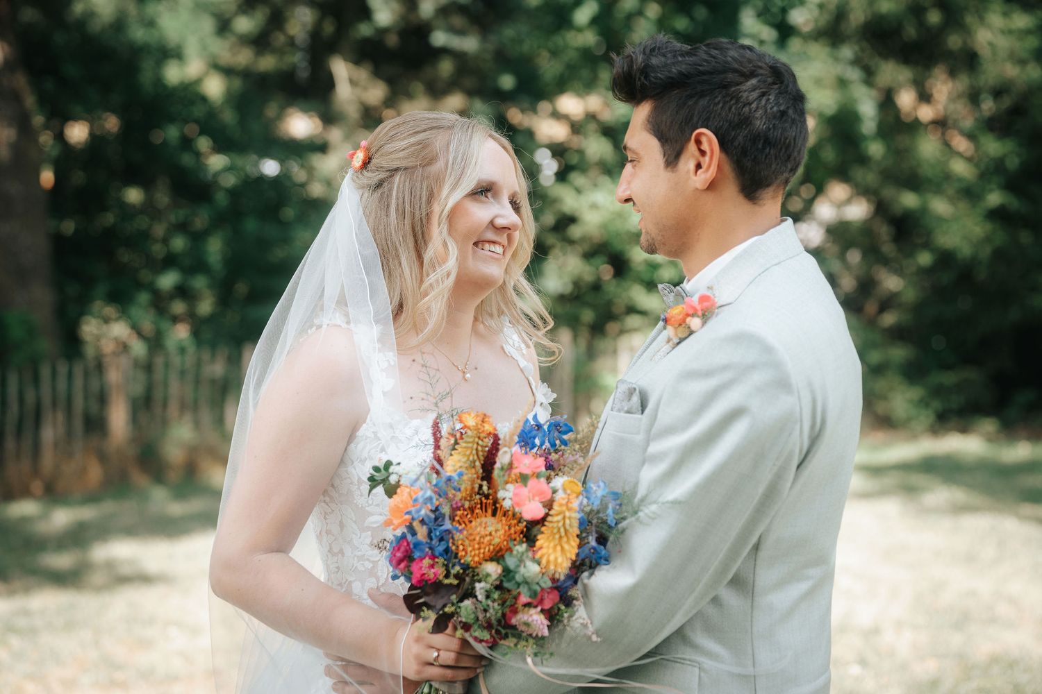 Smiling bride in a lace gown and veil holds a colorful wildflower bouquet while standing beside her groom in a gray suit outdoors.