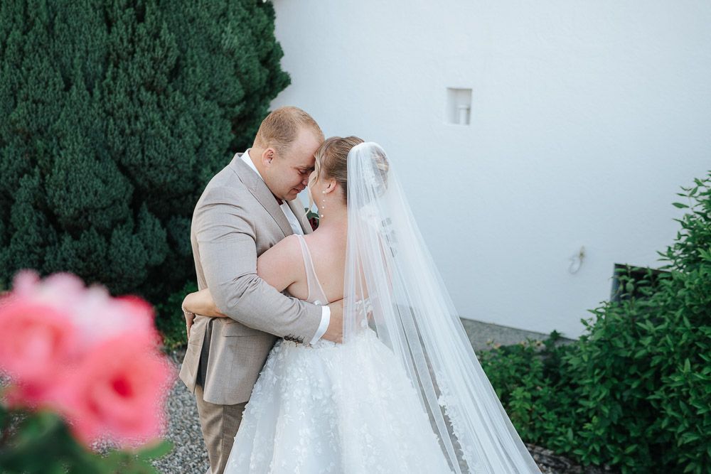 Couple shares an intimate moment in garden setting with pink flowers and greenery on their wedding day.