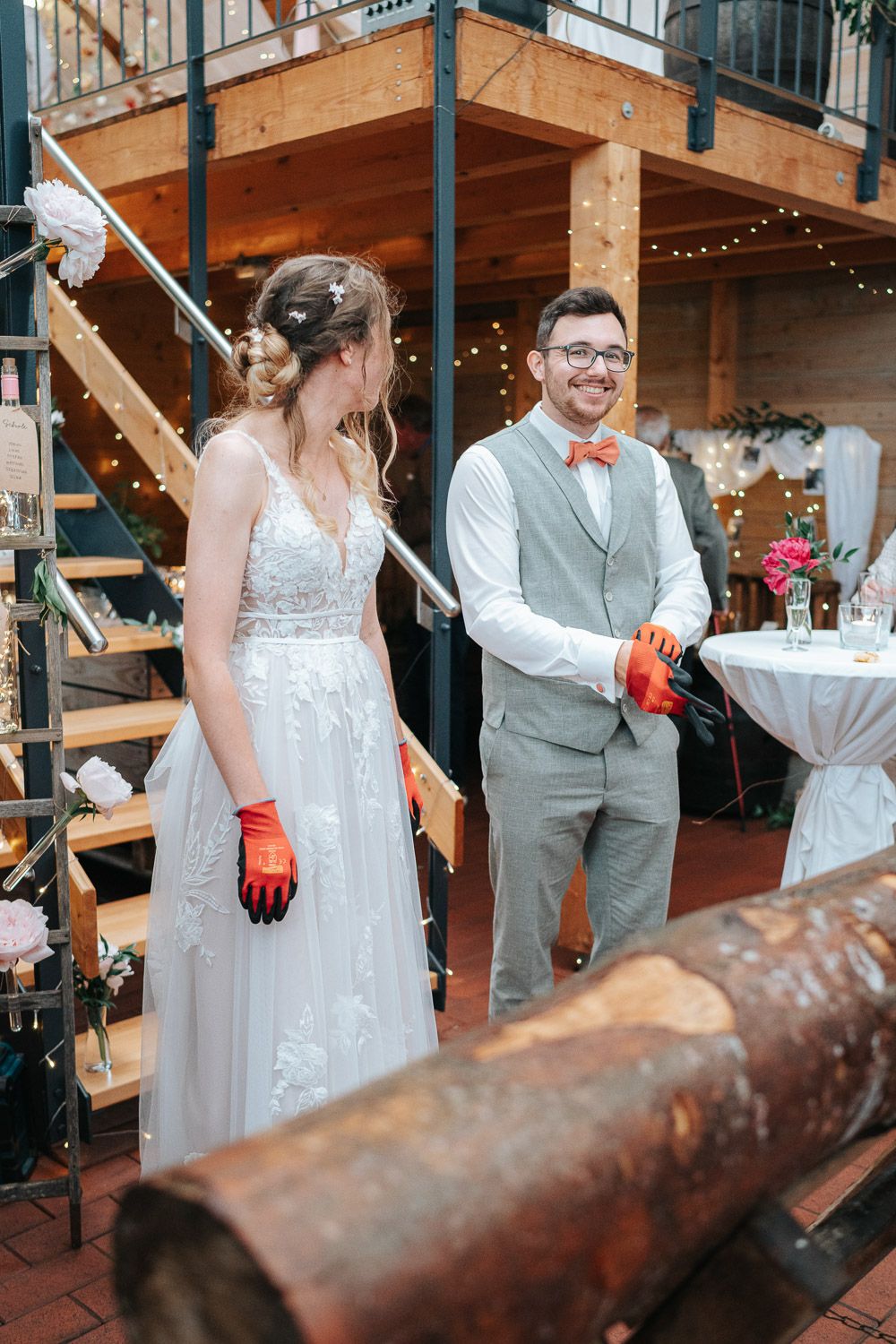 Wedding couple stands with guests viewing a traditional German log cutting ceremony at an outdoor reception.