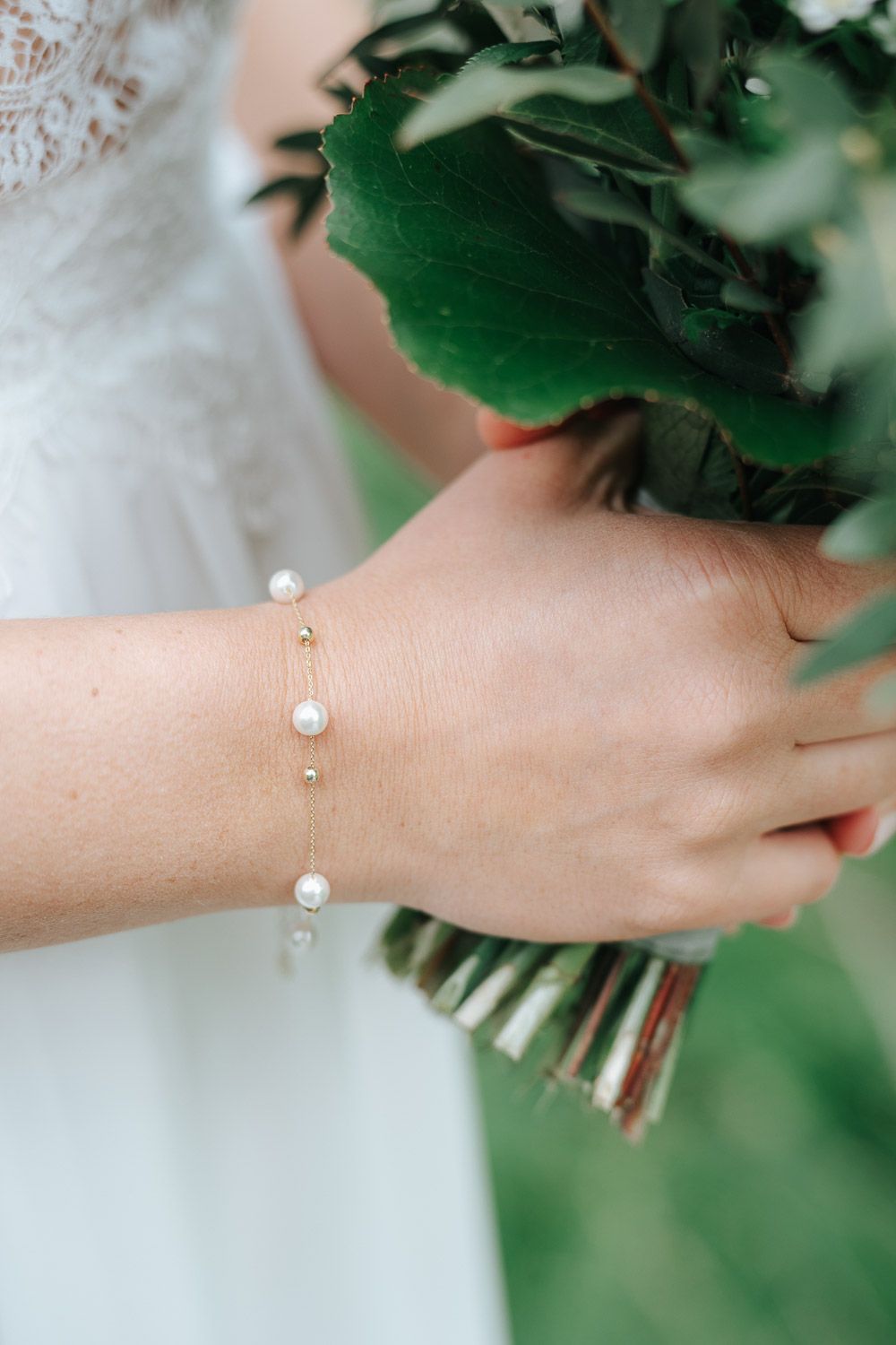 A delicate pearl bracelet on a wrist holding a green floral bouquet with white dress fabric in the background.