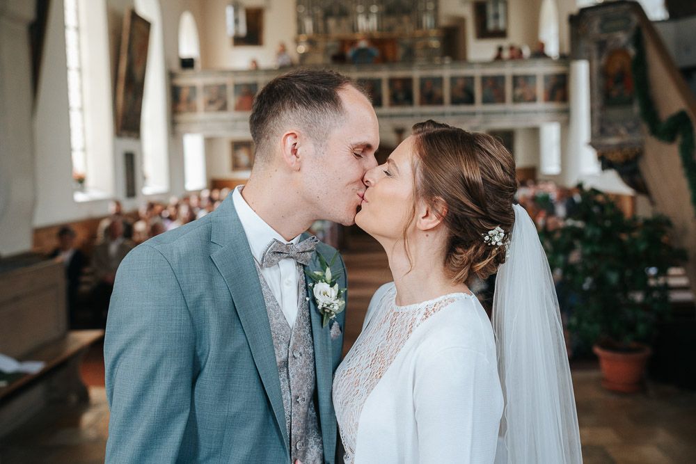 A romantic wedding kiss inside a beautiful traditional church with white walls and wooden pews.