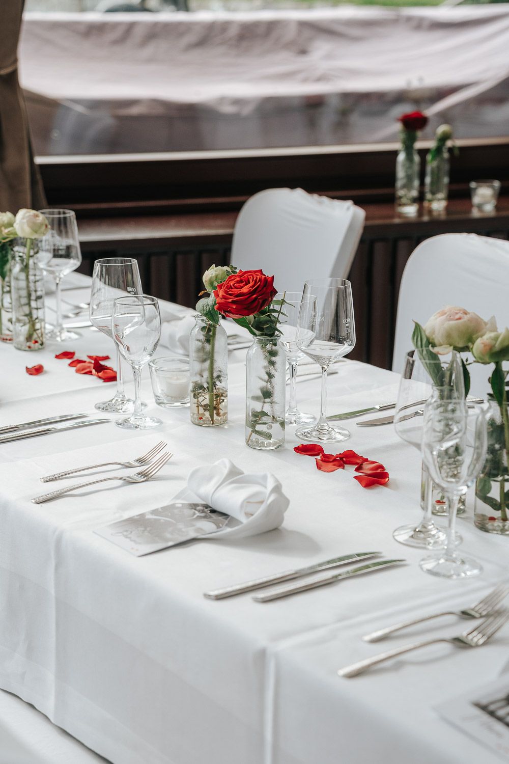 Elegant white table settings at a formal event with red rose petals scattered and wine glasses arranged neatly.
