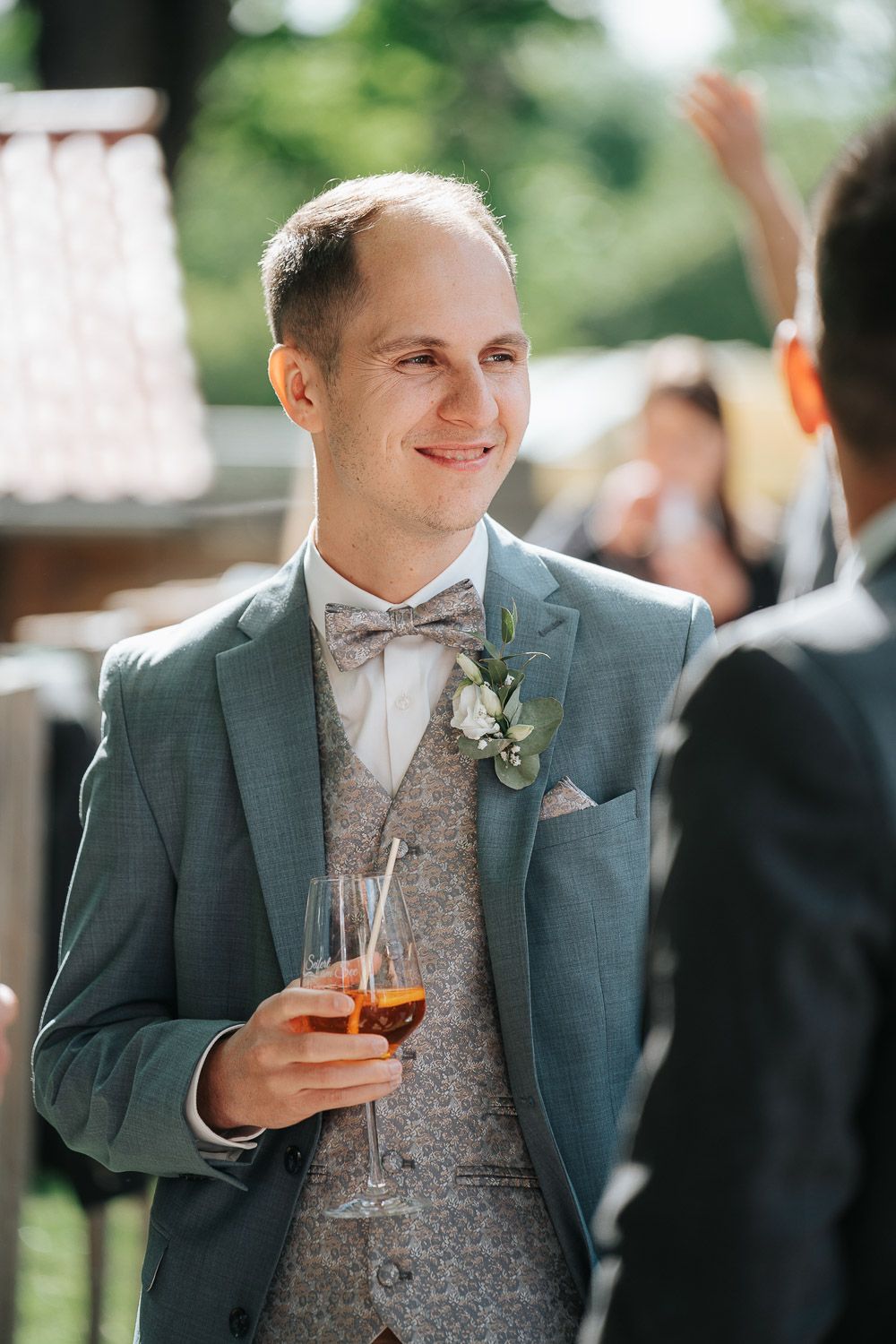 Party guest in gray suit and paisley vest holds cocktail while mingling at outdoor celebration.