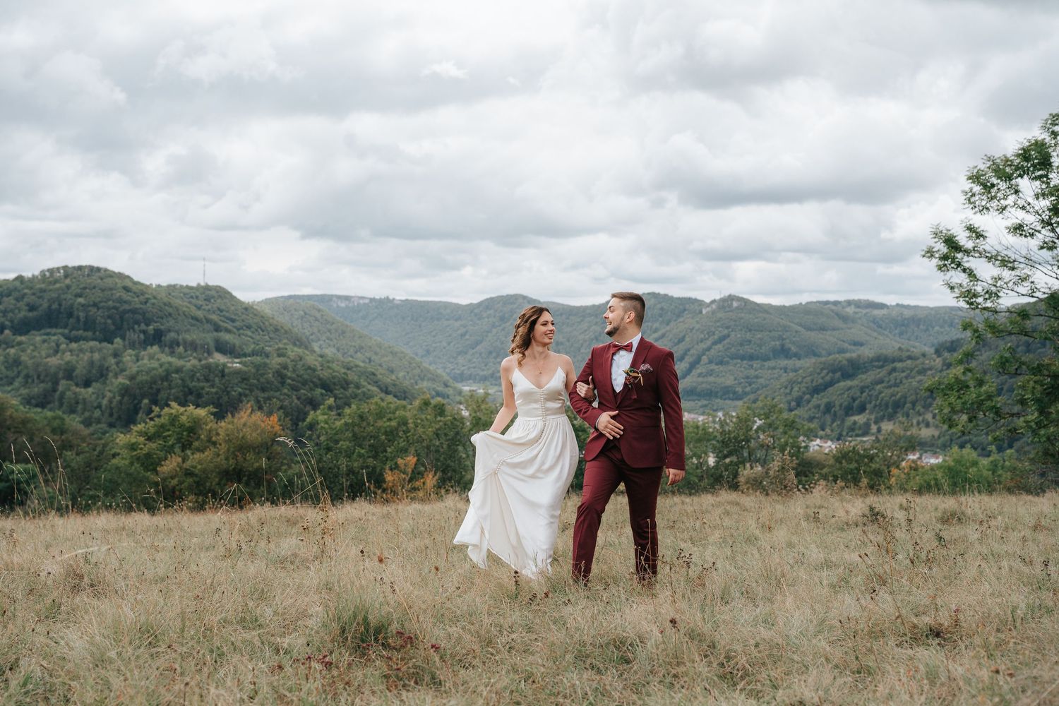 A couple in wedding attire stands together on a hilltop overlooking a scenic mountain landscape on a cloudy day.