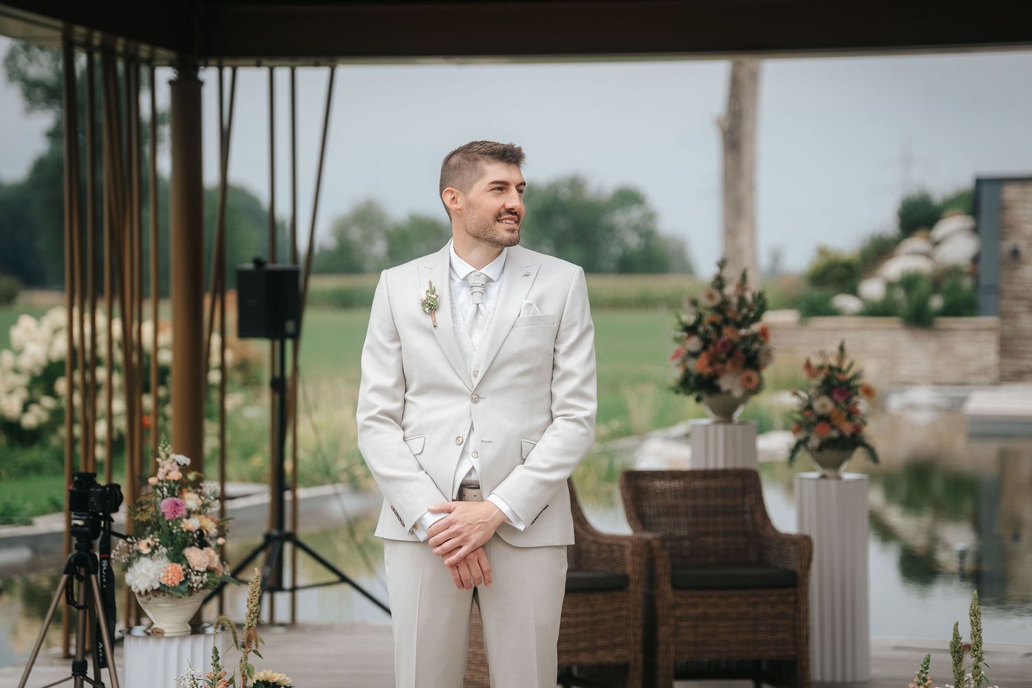 Smiling groom in a light gray suit stands with hands clasped at an outdoor wedding ceremony beside floral arrangements and wooden chairs.