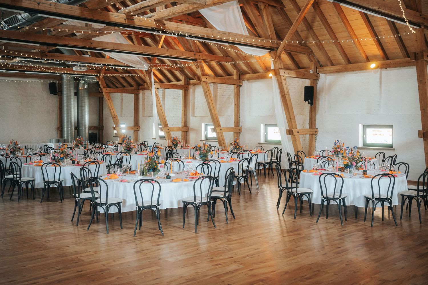 Rustic event hall with exposed wooden beams, round tables with floral centerpieces, black bentwood chairs, and a polished hardwood floor.