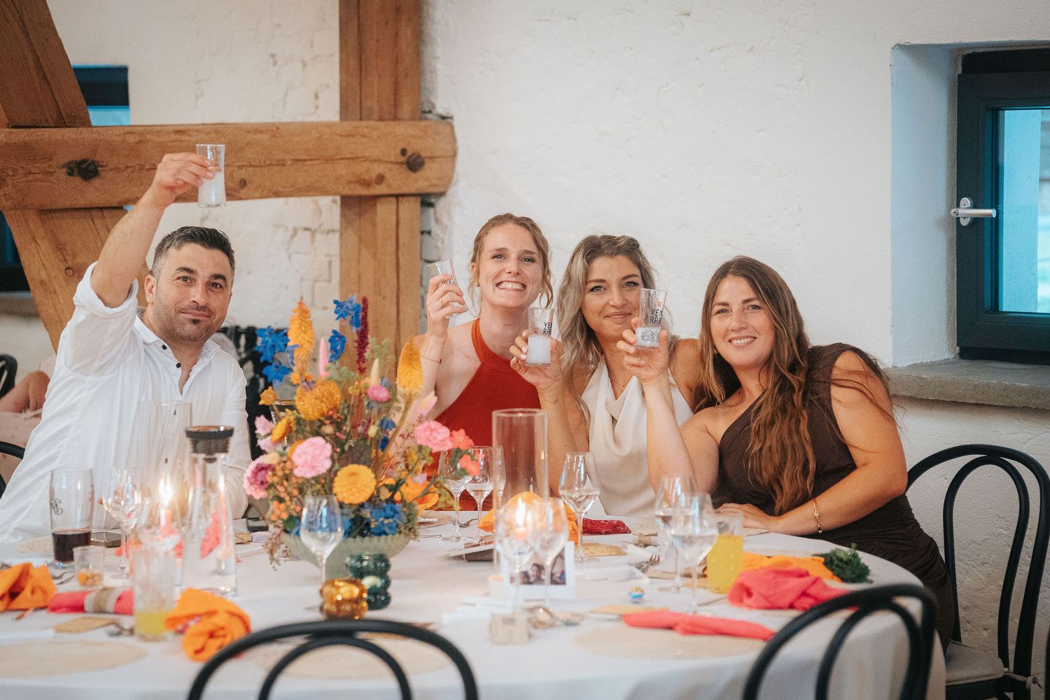 Four smiling guests raise glasses in a toast at a candlelit dining table decorated with colorful flowers in a rustic venue.