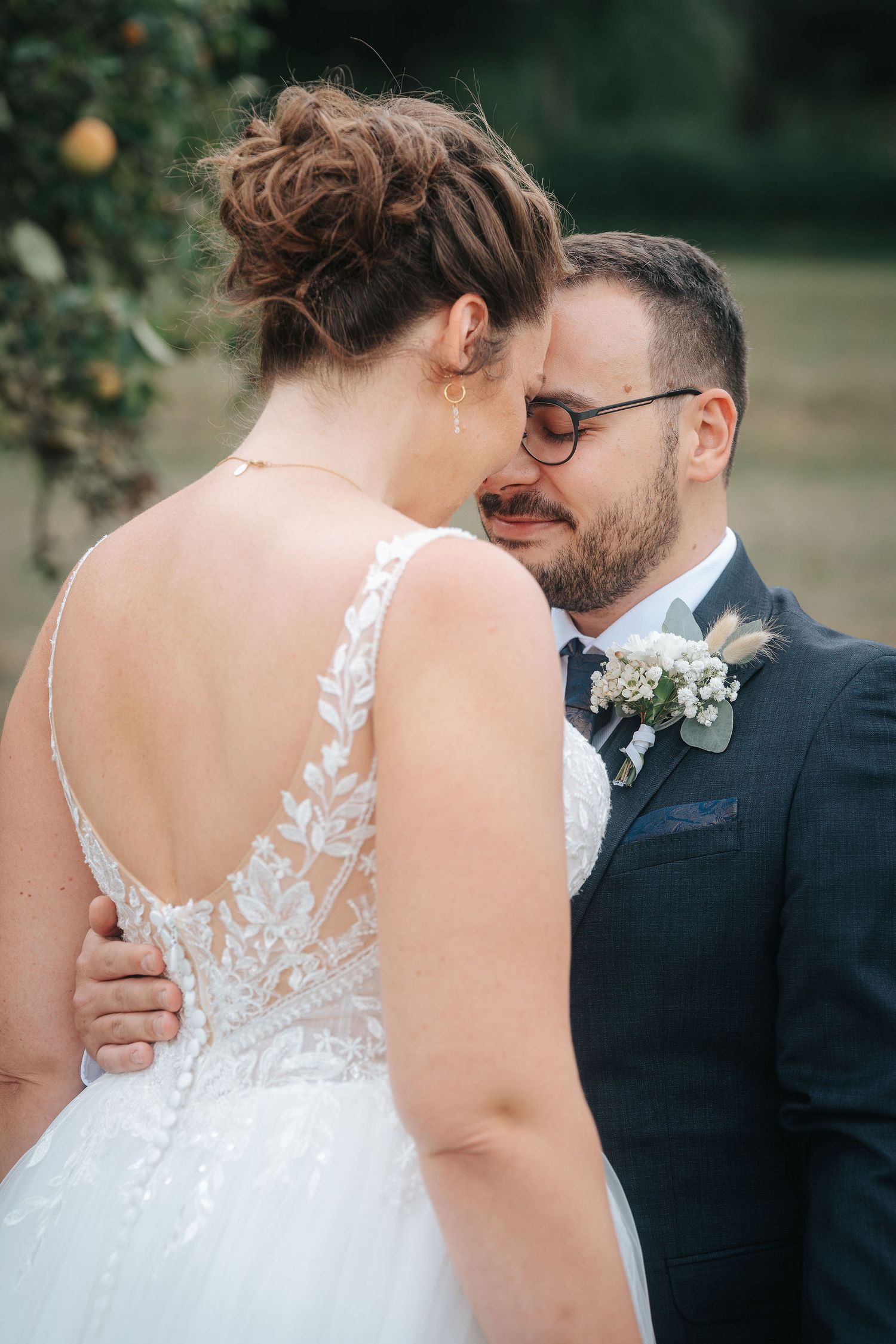 Bride in a lace low-back wedding gown embraces a bearded groom in glasses and a navy suit with a floral boutonniere, foreheads touching.