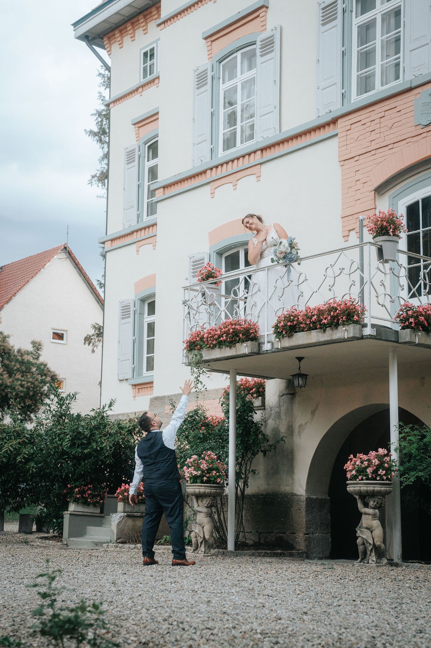 Bride in a white gown leans from a flower-filled balcony toward the groom reaching up from the gravel courtyard of a historic villa.