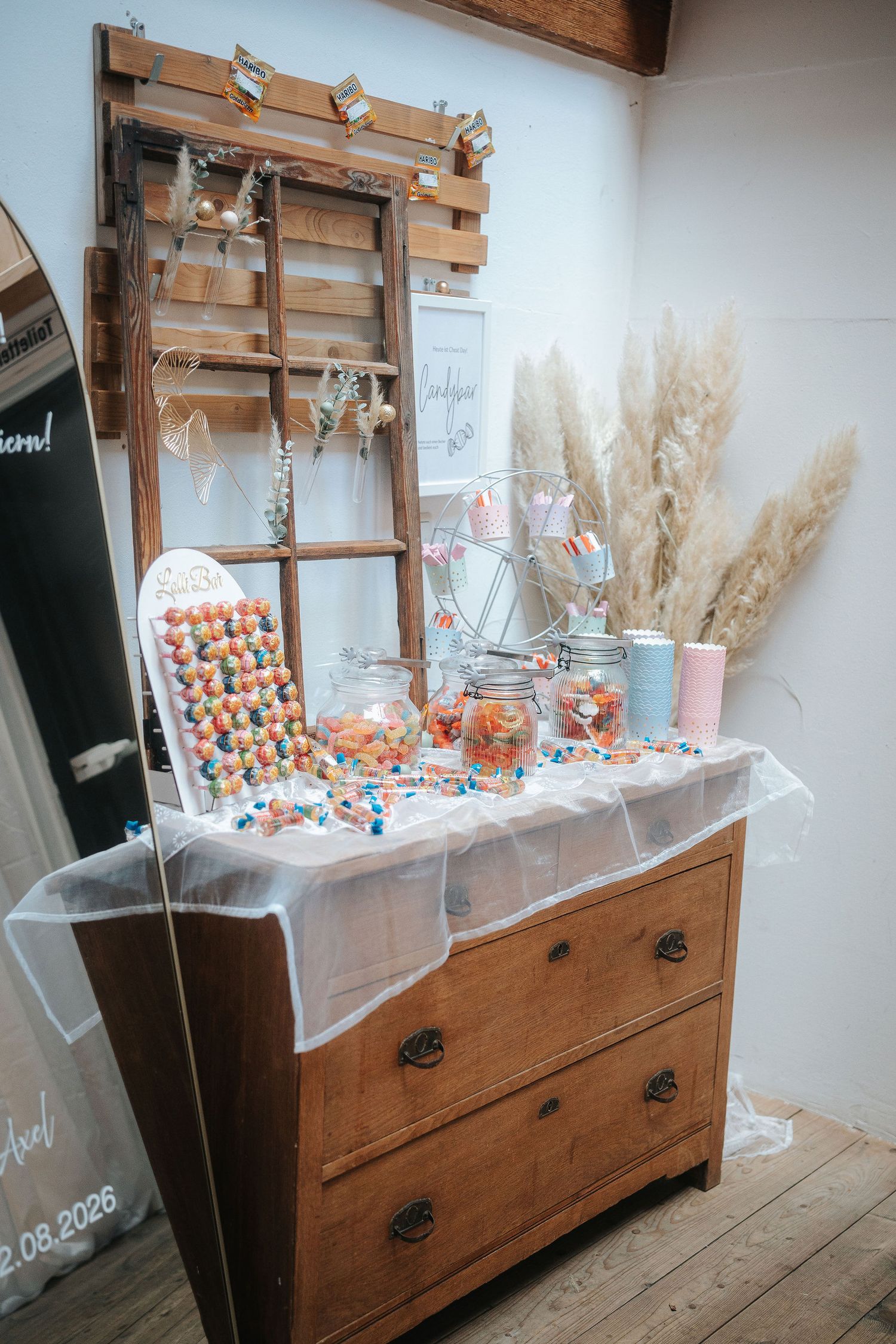 Rustic wooden dresser styled as a wedding candy bar with glass jars, colorful sweets, pampas grass and a wooden ladder decoration.
