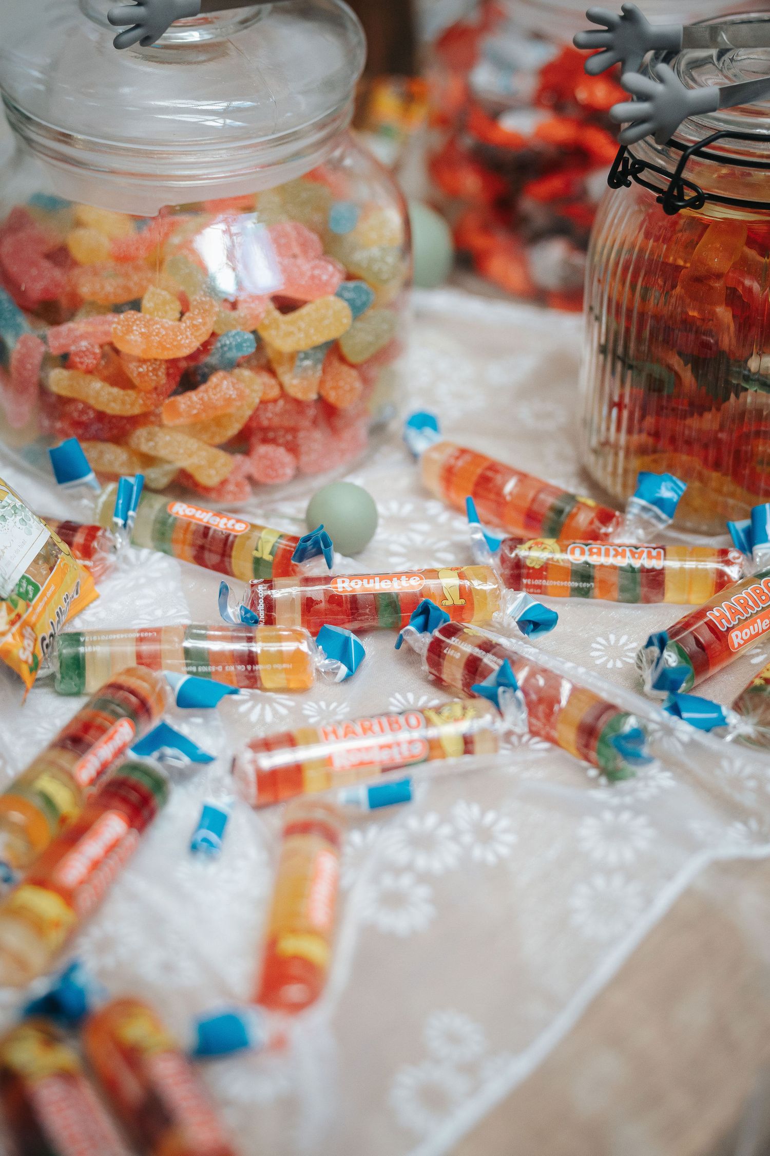Colorful candy rolls in clear wrappers with blue ends scattered on a white lace tablecloth beside glass jars of gummy candies.