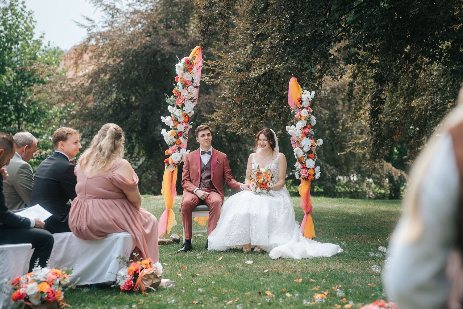 Bride and groom sit together on chairs during an outdoor garden wedding ceremony beneath colorful floral arches as guests look on.