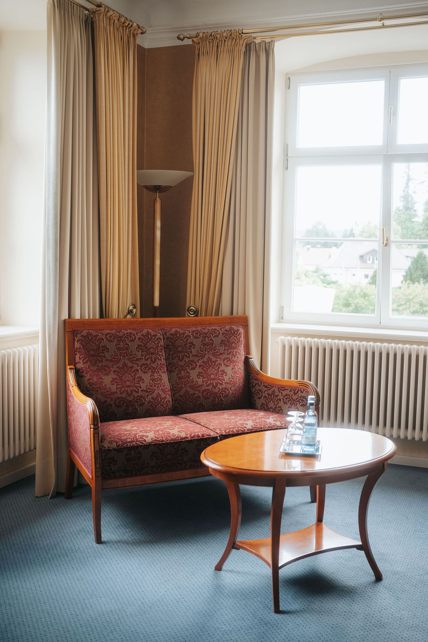 Cozy hotel room corner with a red patterned wooden loveseat, round wooden side table with water bottle, floor lamp, and a bright window.