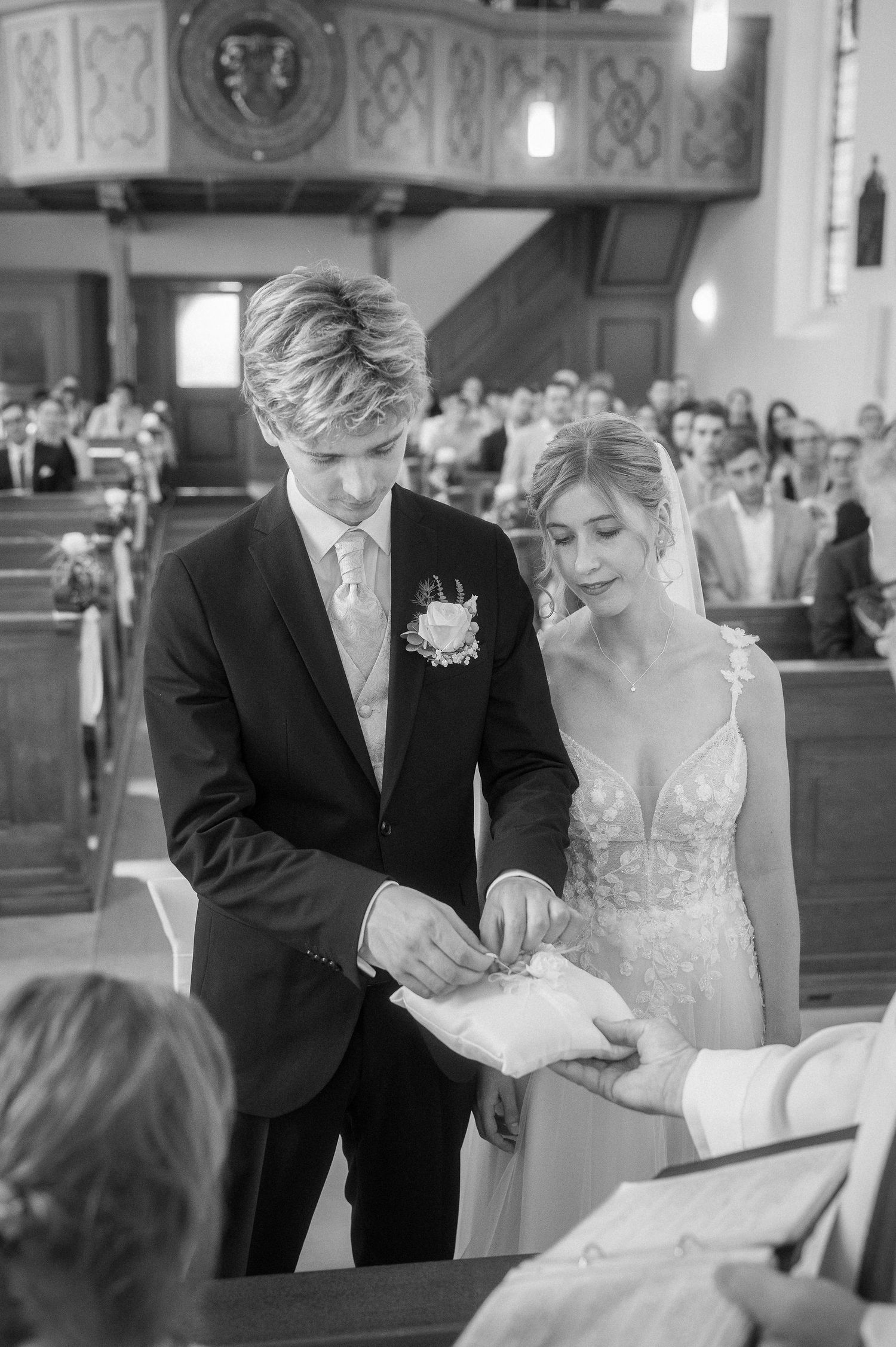 Black and white photo of a bride and groom exchanging rings during a church wedding ceremony as guests watch from the pews.