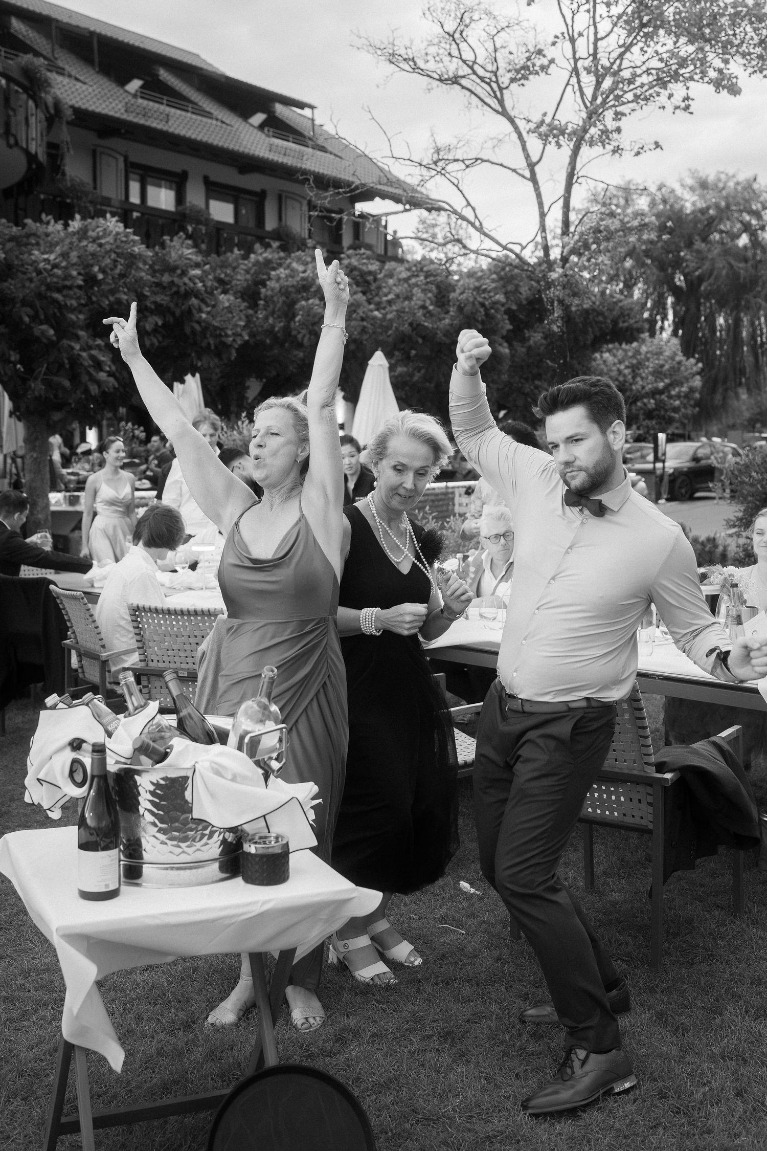 Two women and a bearded man in a bow tie dance joyfully with raised arms at an outdoor garden wedding reception.