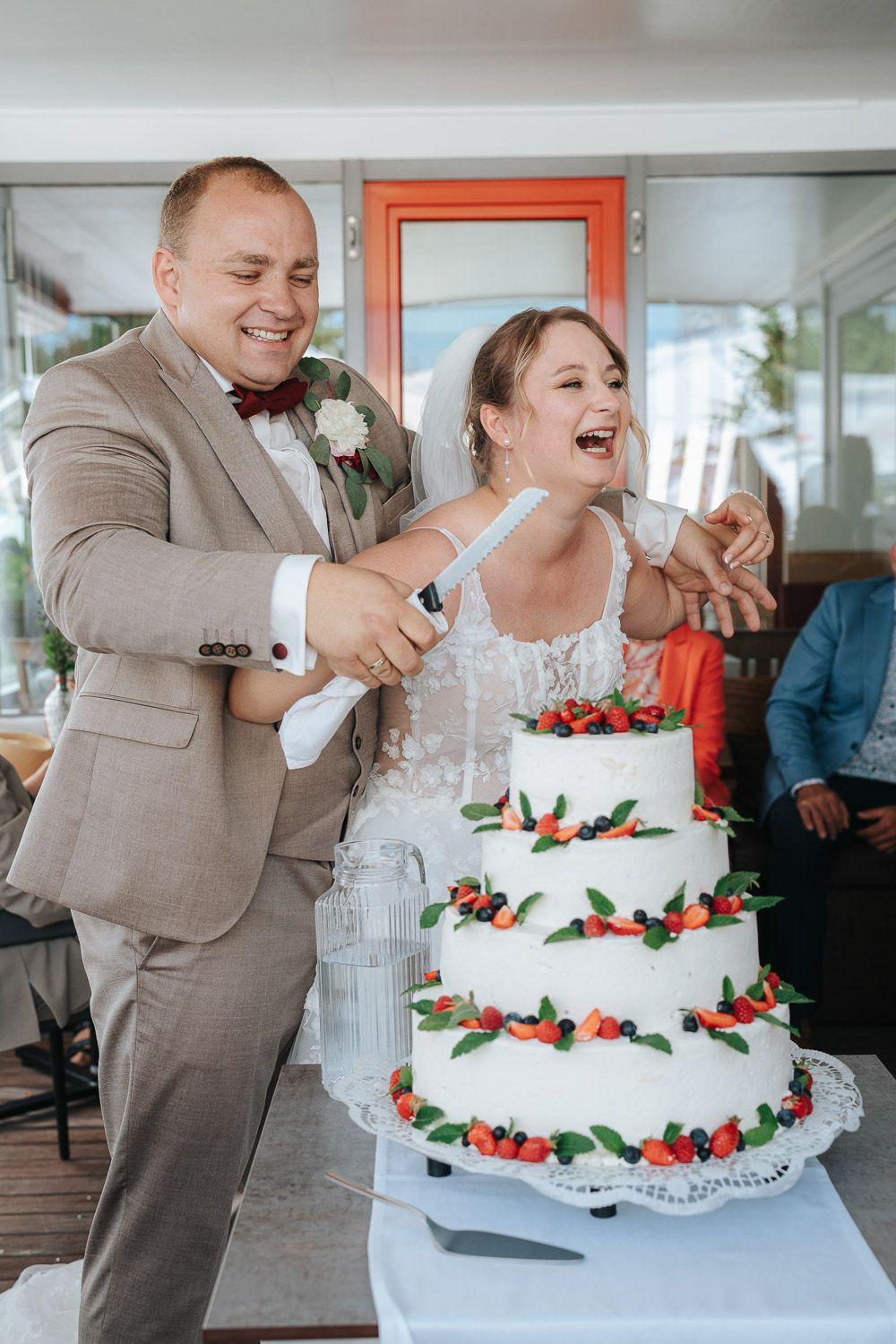 Wedding couple laughs together while cutting their white tiered cake decorated with fresh berries in panoramic sequence.