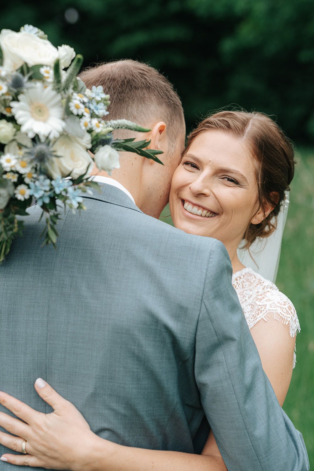 A joyful wedding moment captured outdoors with lace details, gray suit and white floral bouquet with blue accents.