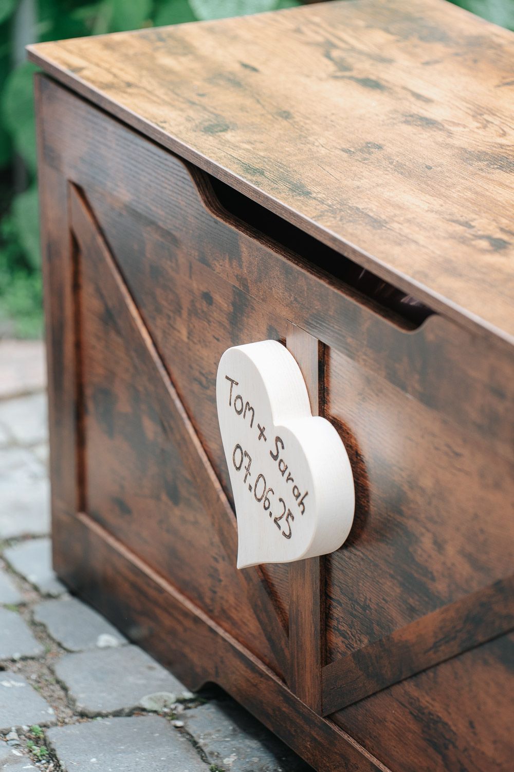 Rustic wooden wedding card box with heart-shaped detail and handwritten text sitting on stone pavement.