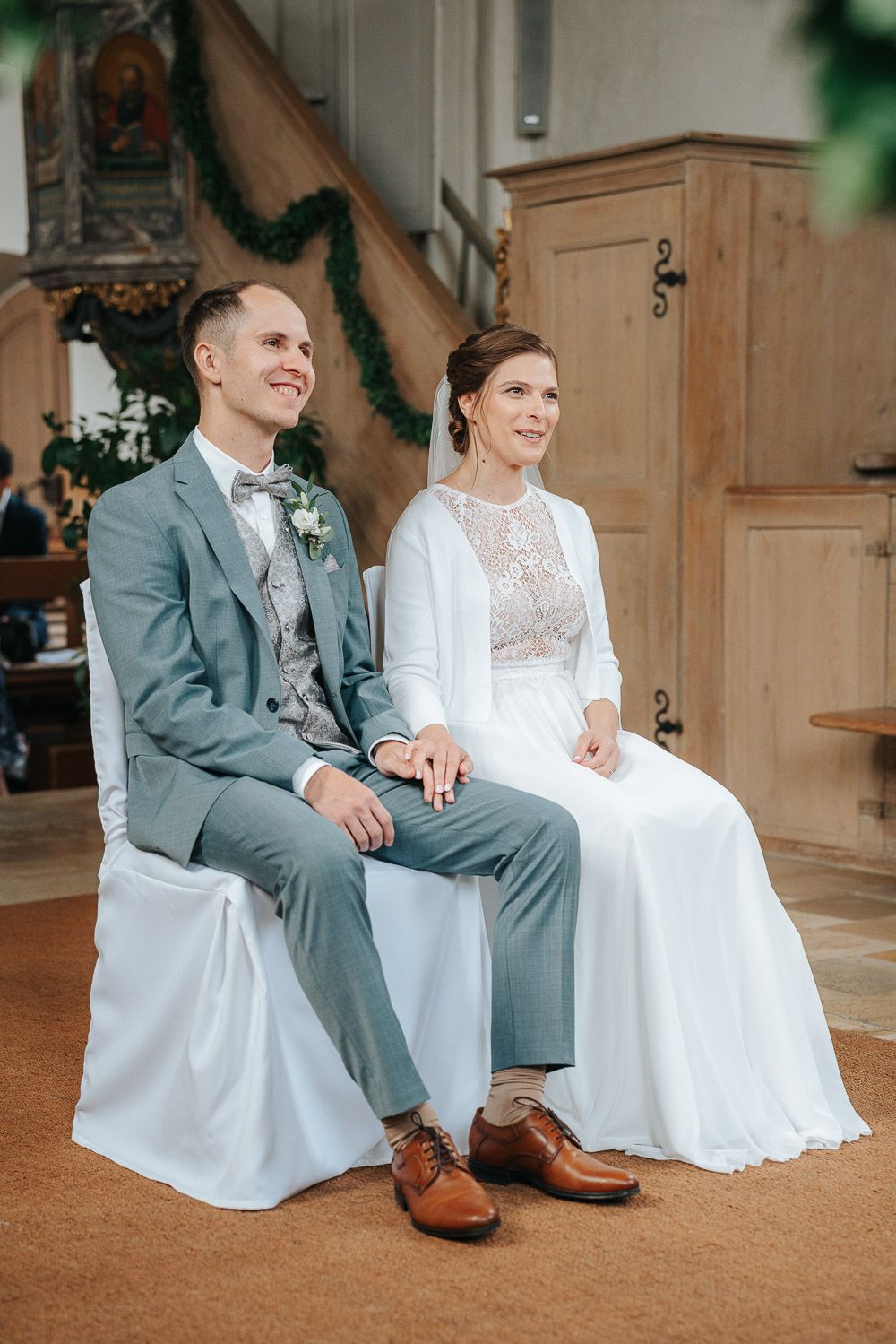 A couple in wedding attire sits on chairs during their ceremony, with the bride wearing a lace gown and groom in gray suit.
