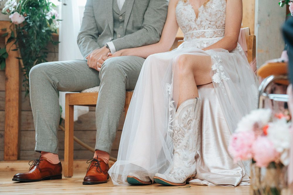 Close-up of a couple sitting on wooden chairs at a wedding, showing detailed lace dress and gray suit with brown shoes.