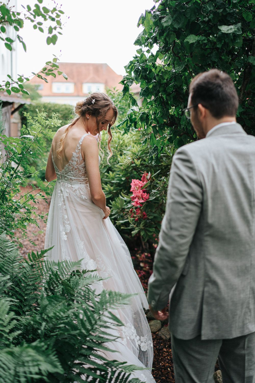 A romantic garden wedding scene with a lace-back gown and grey suit among lush greenery and pink flowers.