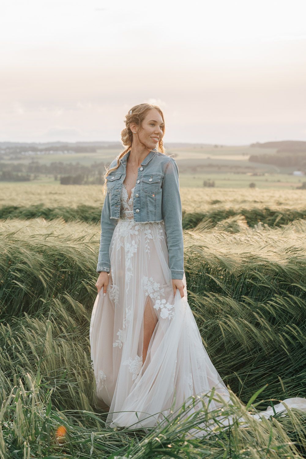 Bride in white wedding dress and denim jacket stands in tall grass field during sunset.