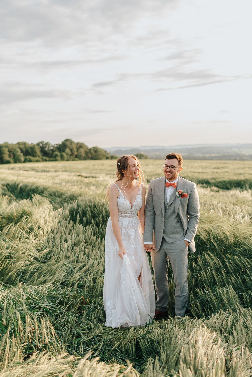 A couple in wedding attire stands together in a scenic field of tall grass during golden hour.