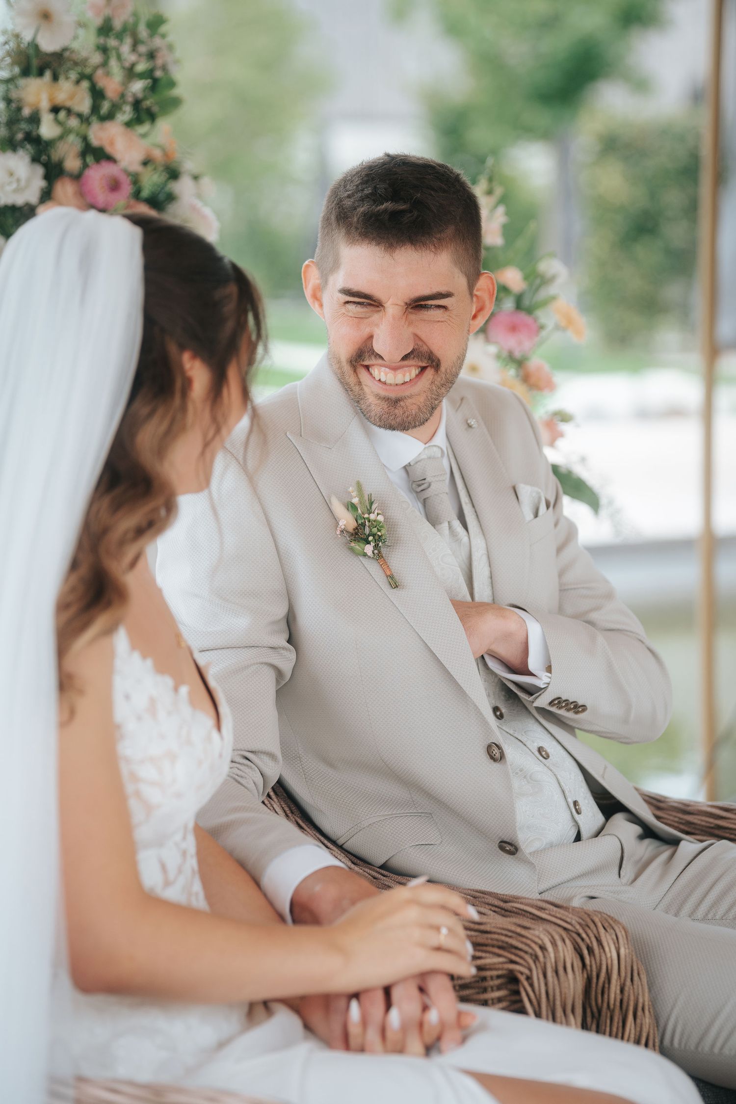 Smiling groom in a beige suit holds hands with a bride in a lace dress and veil during an outdoor wedding ceremony with flowers.