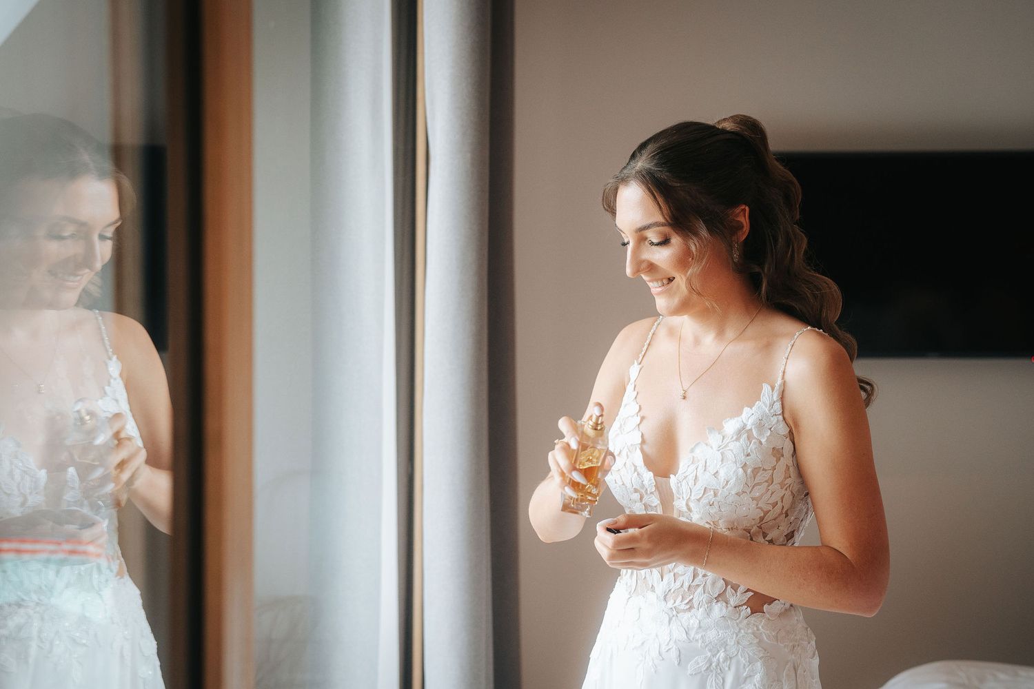 Smiling bride in a white lace wedding dress applying perfume by a window while getting ready, with her reflection visible in the mirror.