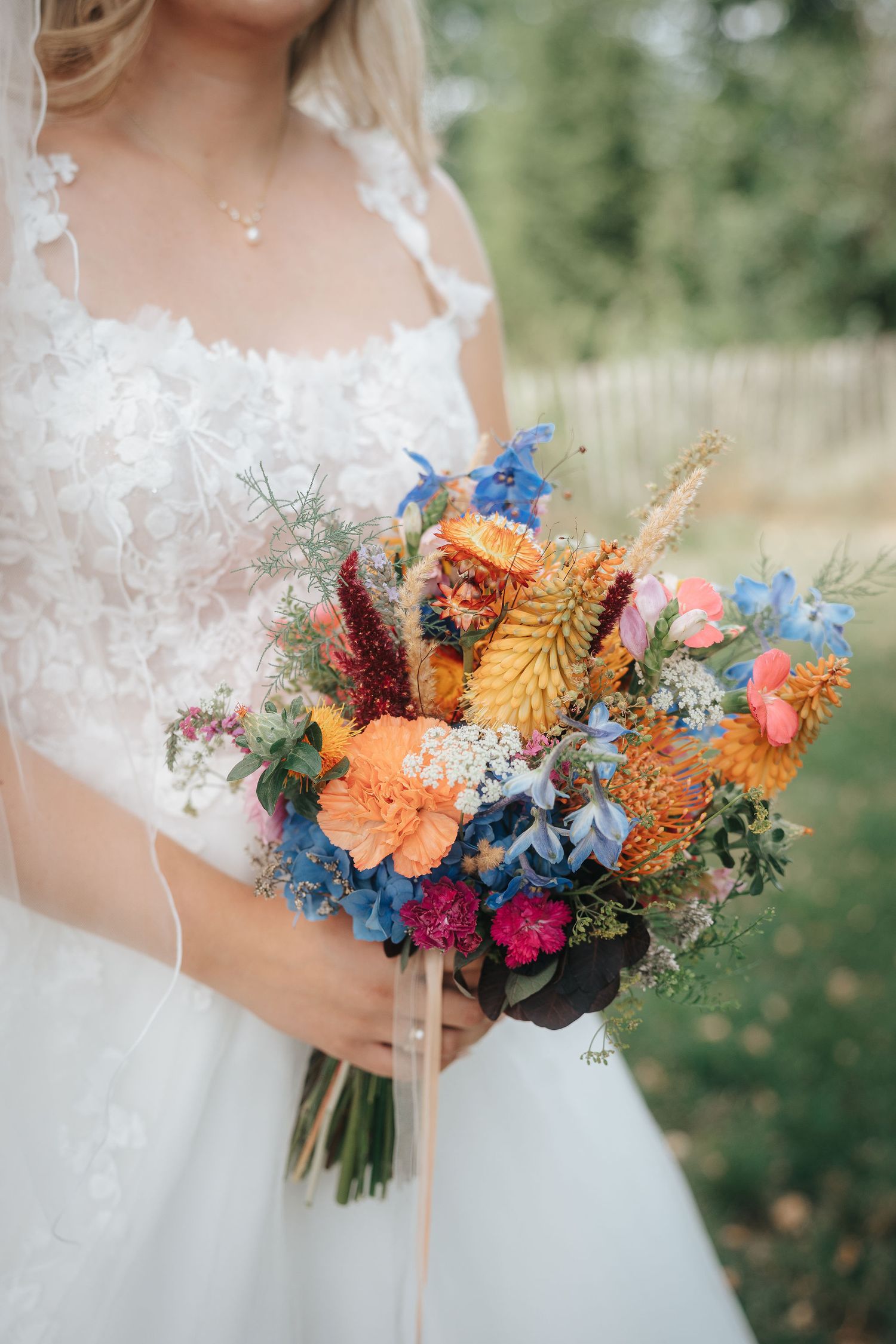 Bride in a white lace gown holds a colorful wildflower bouquet of orange, blue, pink and yellow blooms outdoors.