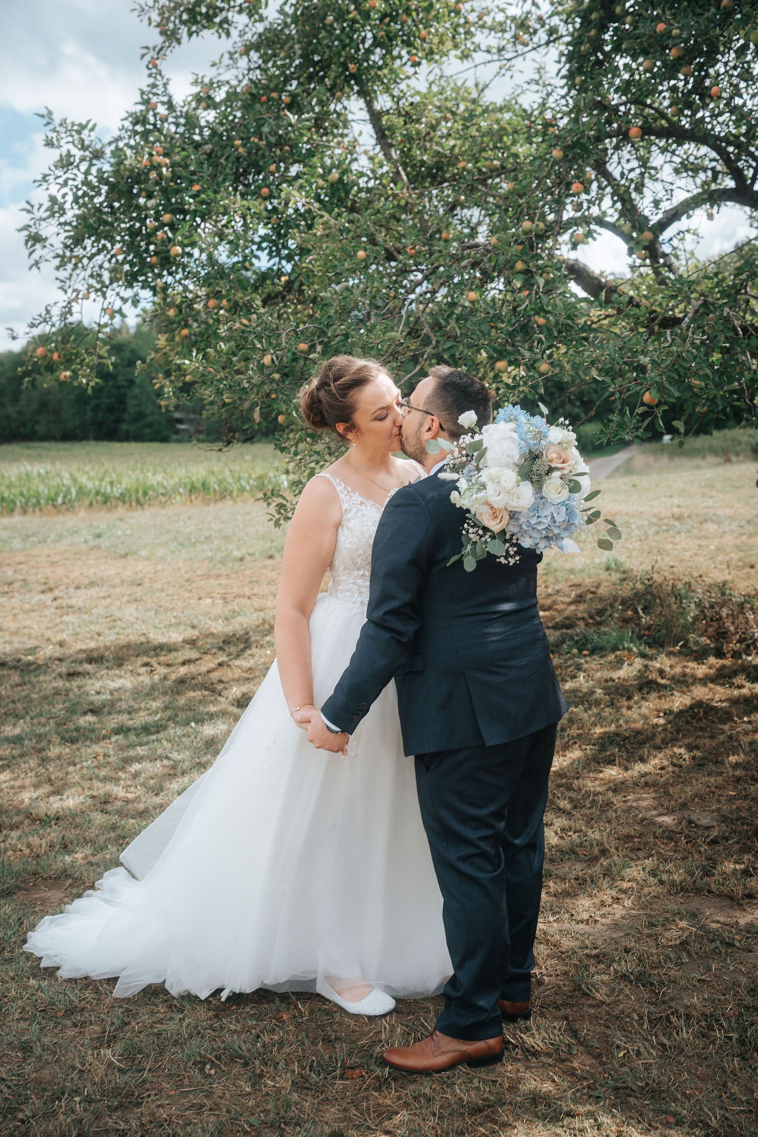 Bride in a white tulle gown and groom in a navy suit share a kiss under an apple tree, holding a blue and white wedding bouquet.