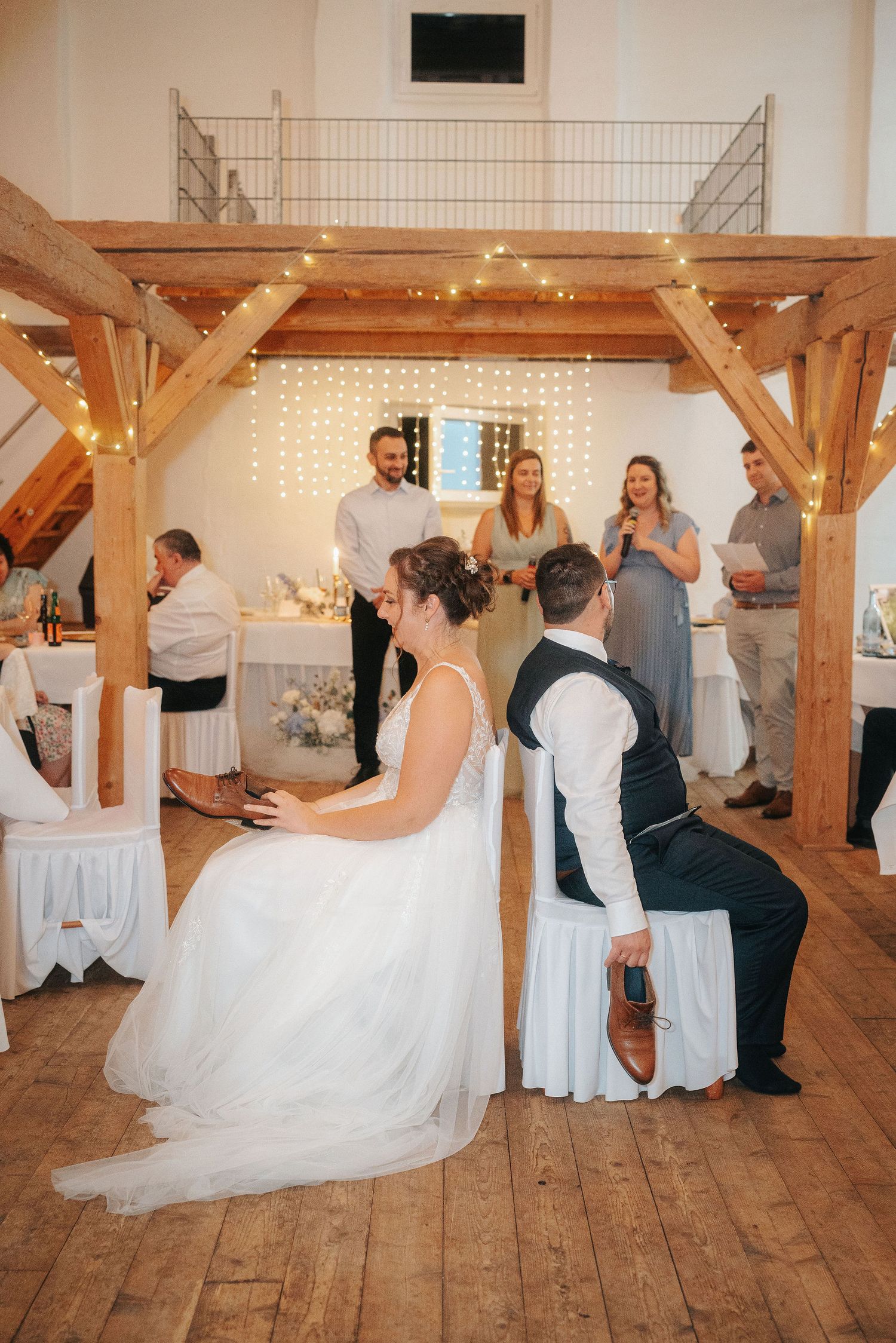 Bride and groom sit back to back holding shoes during a wedding reception game while guests watch beneath a rustic wooden beam loft.