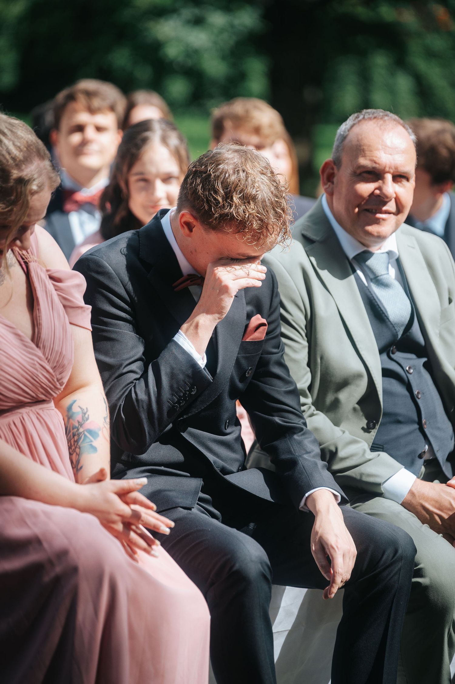 Emotional groom in a dark suit wipes his eyes during an outdoor wedding ceremony while seated guests smile and look on.