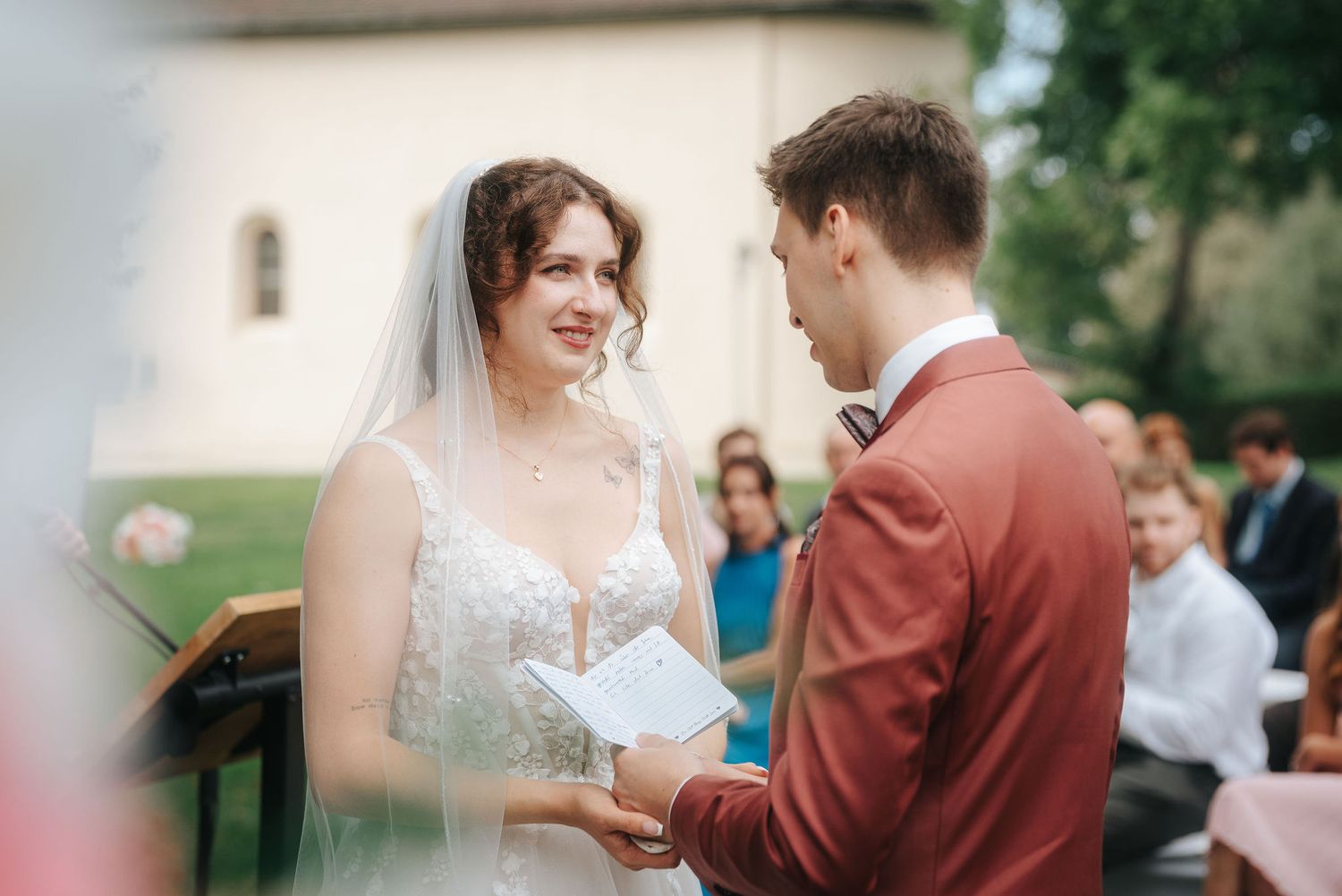 Smiling bride in a lace wedding gown and veil holds a paper while the groom in a burgundy suit reads his vows at an outdoor ceremony.