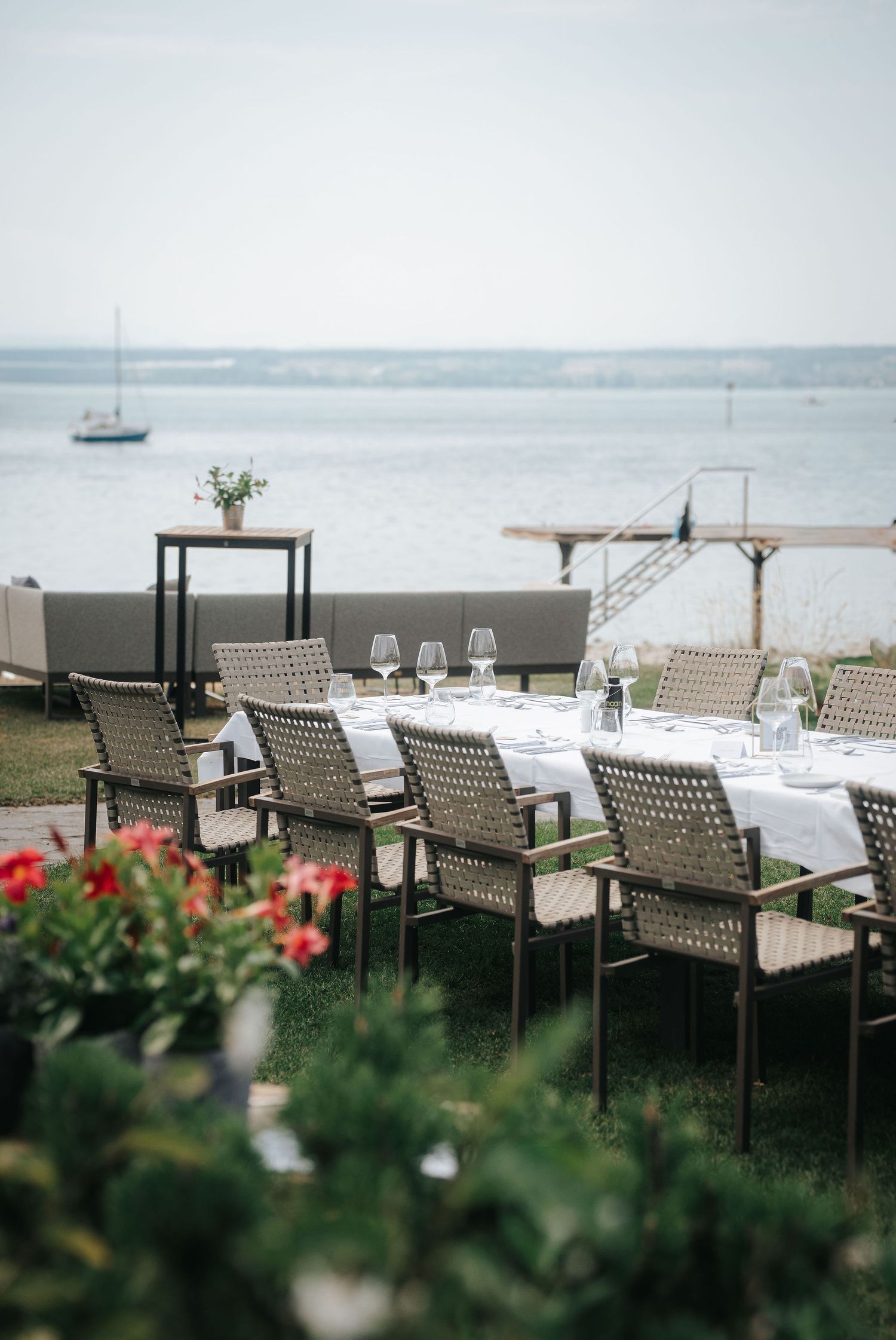 Elegant waterfront dining table with wicker chairs and wine glasses on a lakeside lawn, with red flowers, a dock and a sailboat beyond.