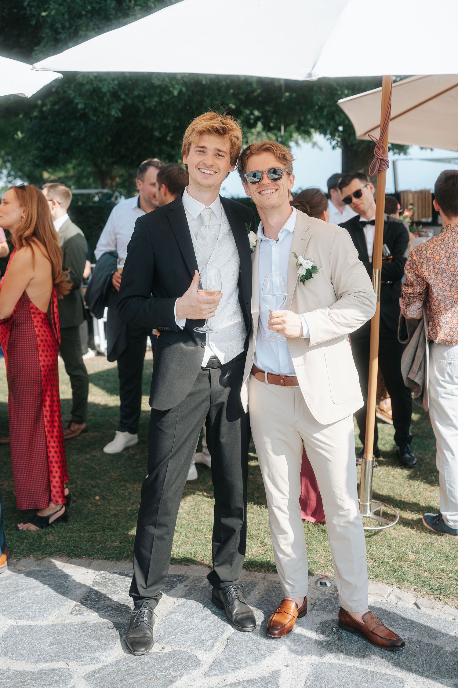 Two smiling young men in suits pose together at an outdoor wedding reception under white umbrellas, holding drinks among guests.