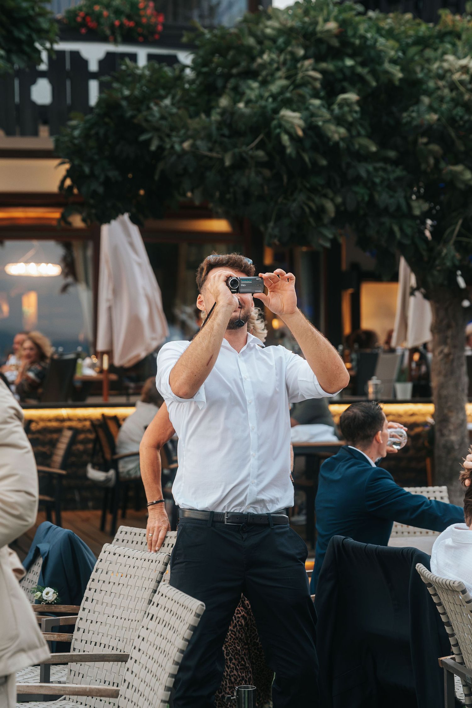 Man in a white shirt and dark trousers raises a small camera to take a photo at a busy outdoor café terrace with wicker chairs.