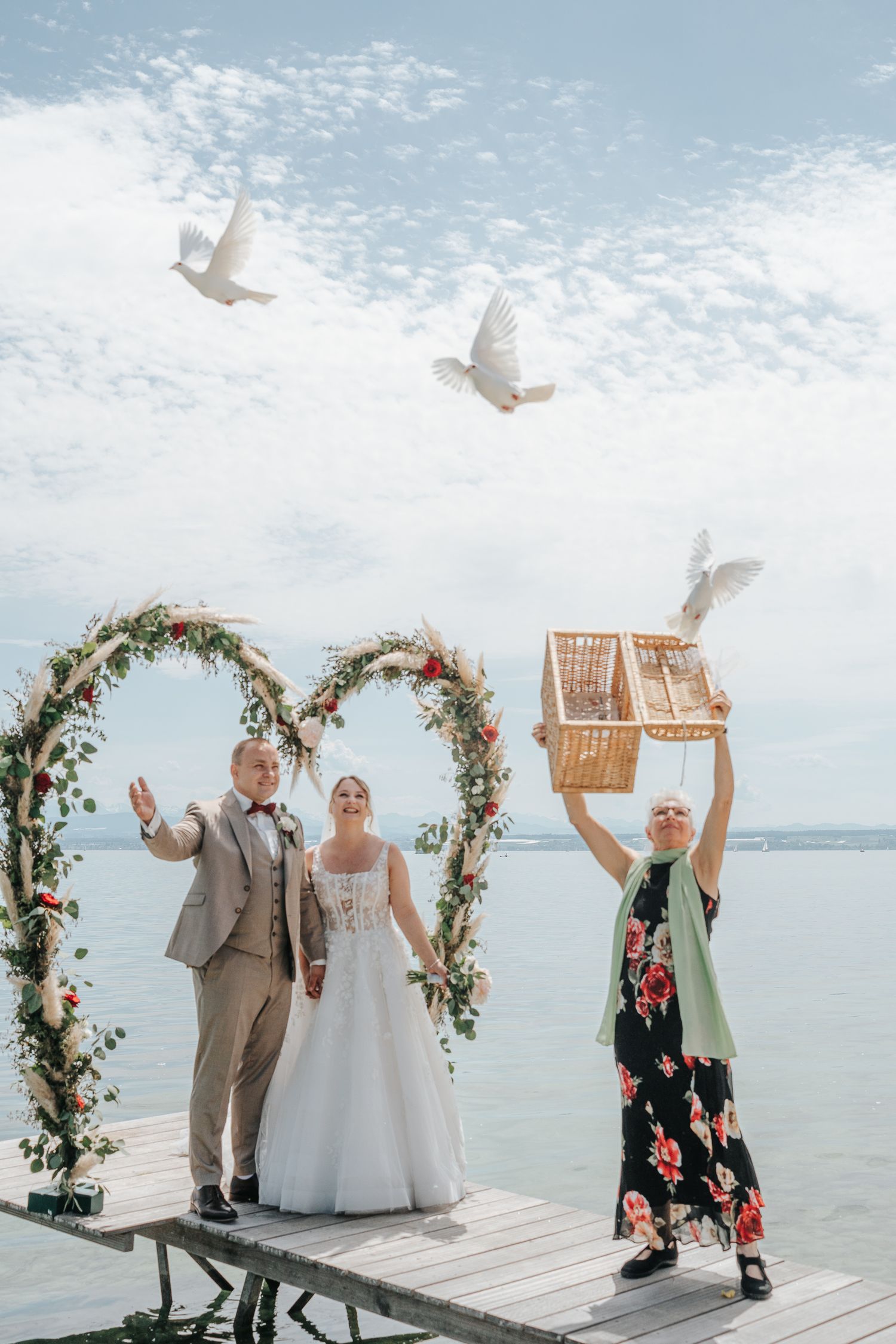 A couple celebrates on a waterfront dock with floral arch while someone releases white doves from a cage.
