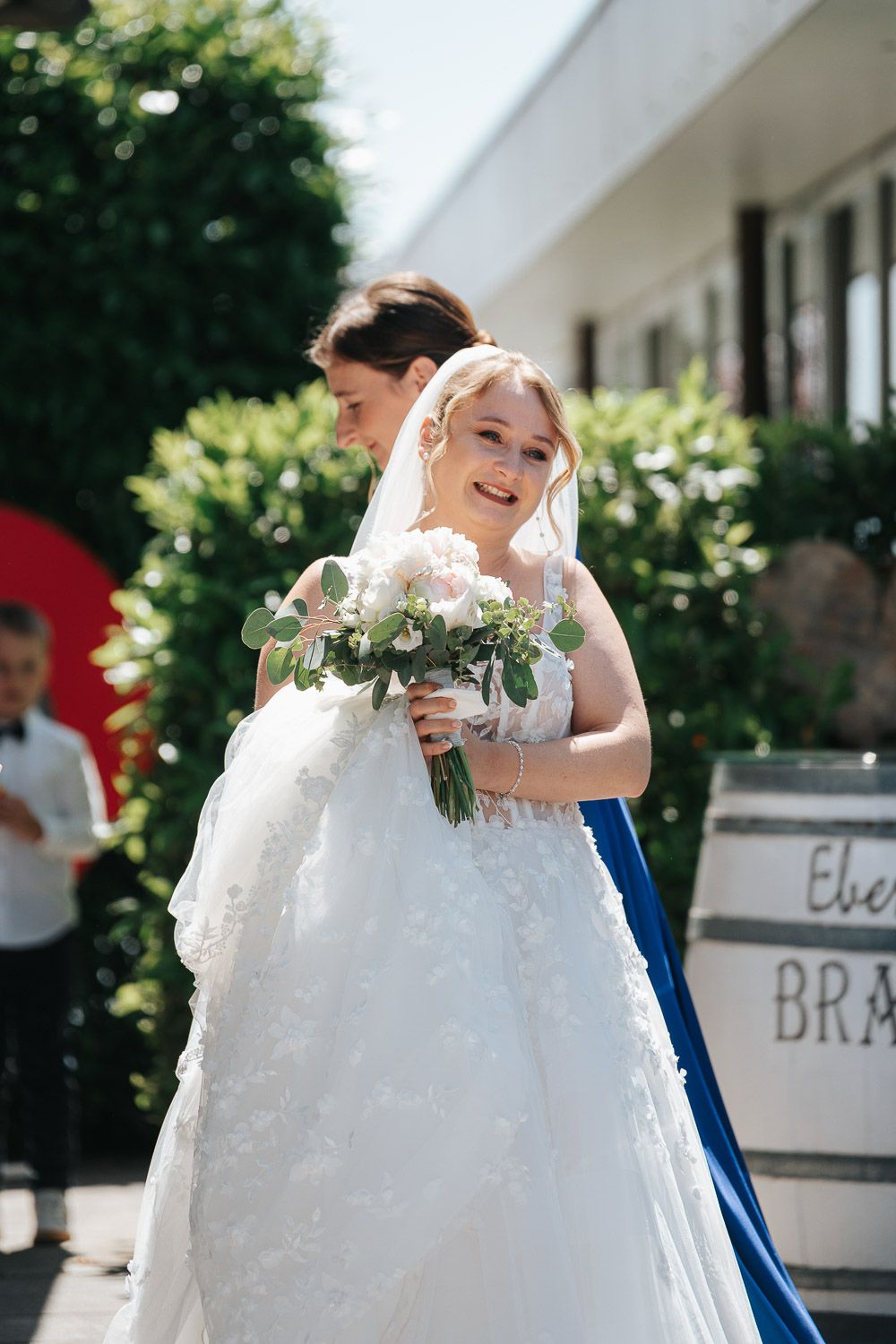 A bride in a beautiful white lace gown holds a pale bouquet while standing outdoors on a sunny day.