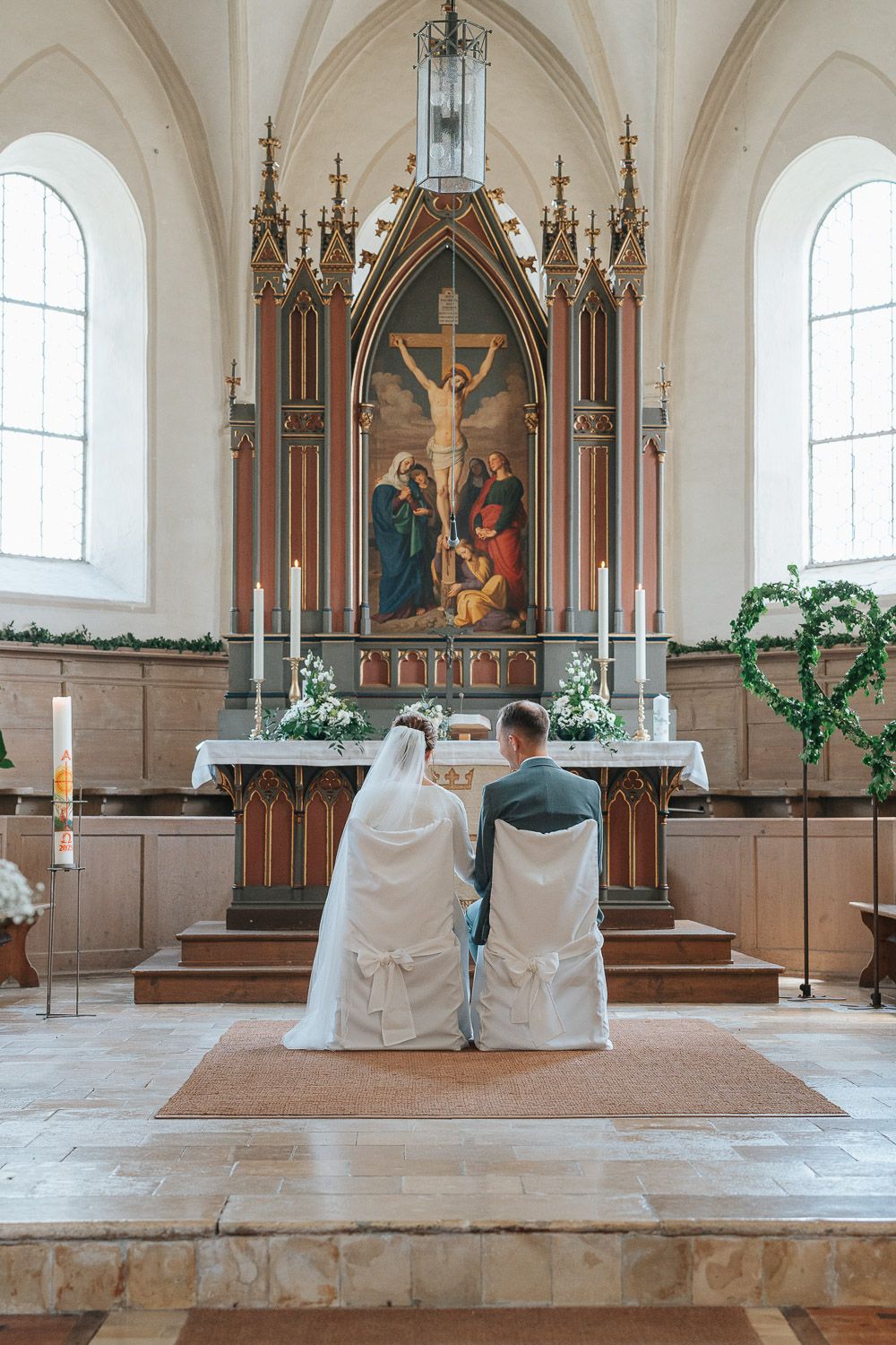 Two people in white cloaks sit before an ornate Gothic church altar with a crucifixion painting and stained glass windows.