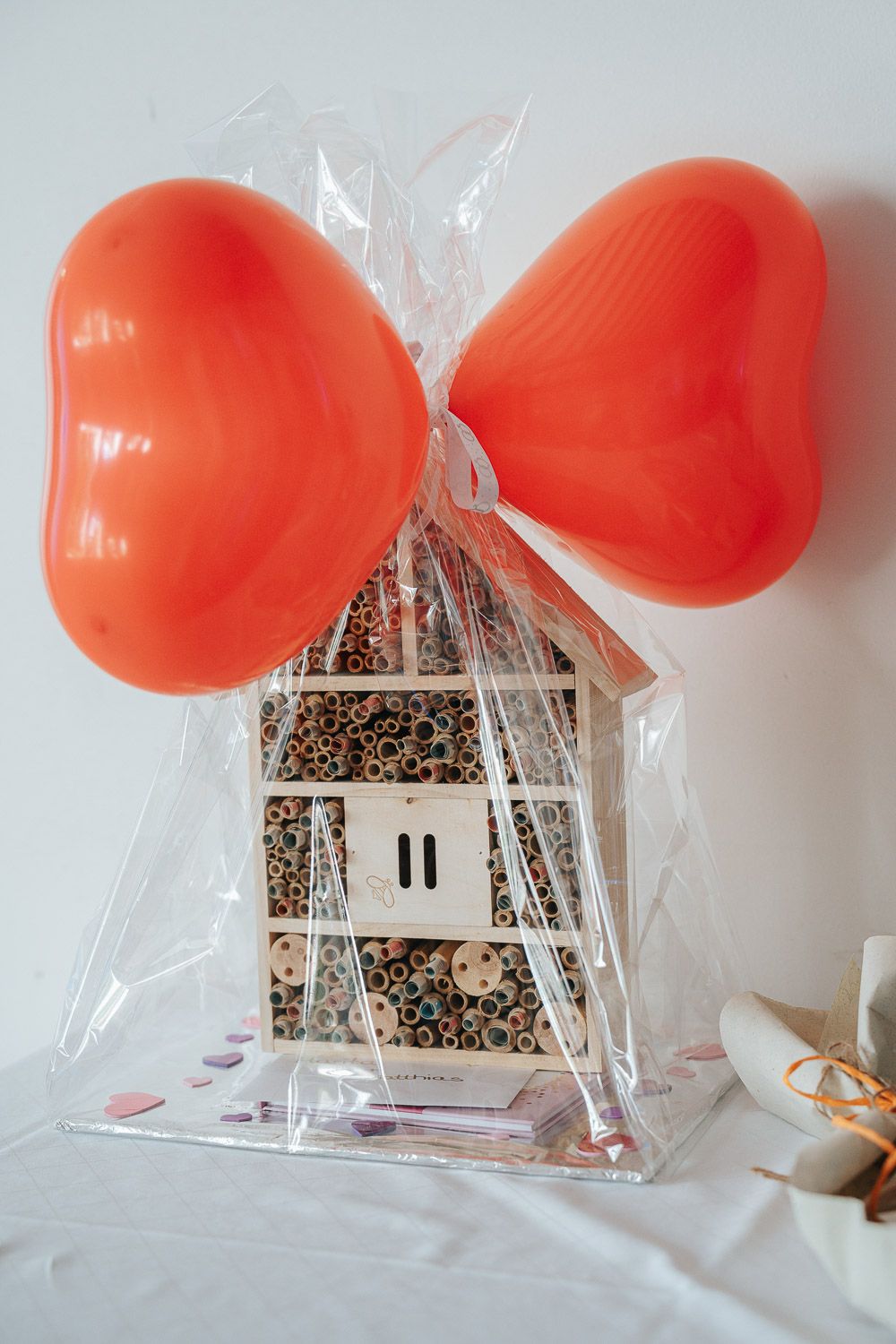 Small clear gift box filled with seeds or grains decorated with two red heart balloons tied with clear plastic ribbon.
