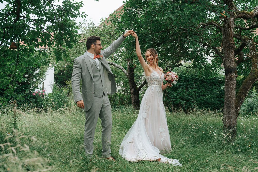 A wedding couple dances joyfully in a lush green forest setting during an outdoor summer celebration.