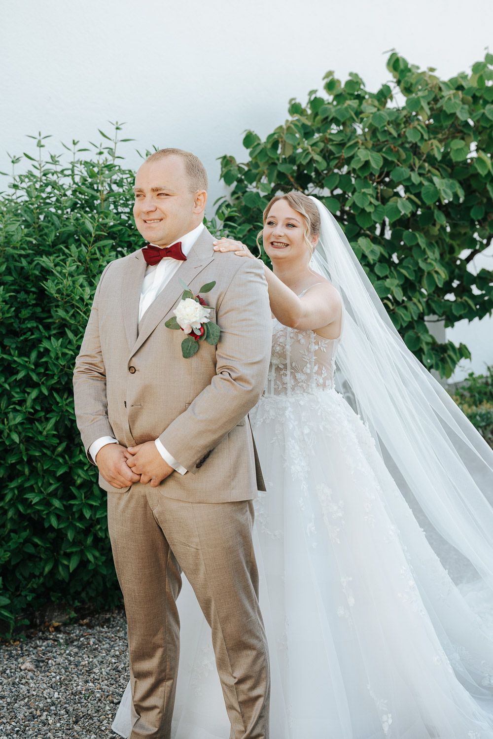 A happy couple shares joyous laughter in a garden on their wedding day wearing tan suits and white gown with veil.