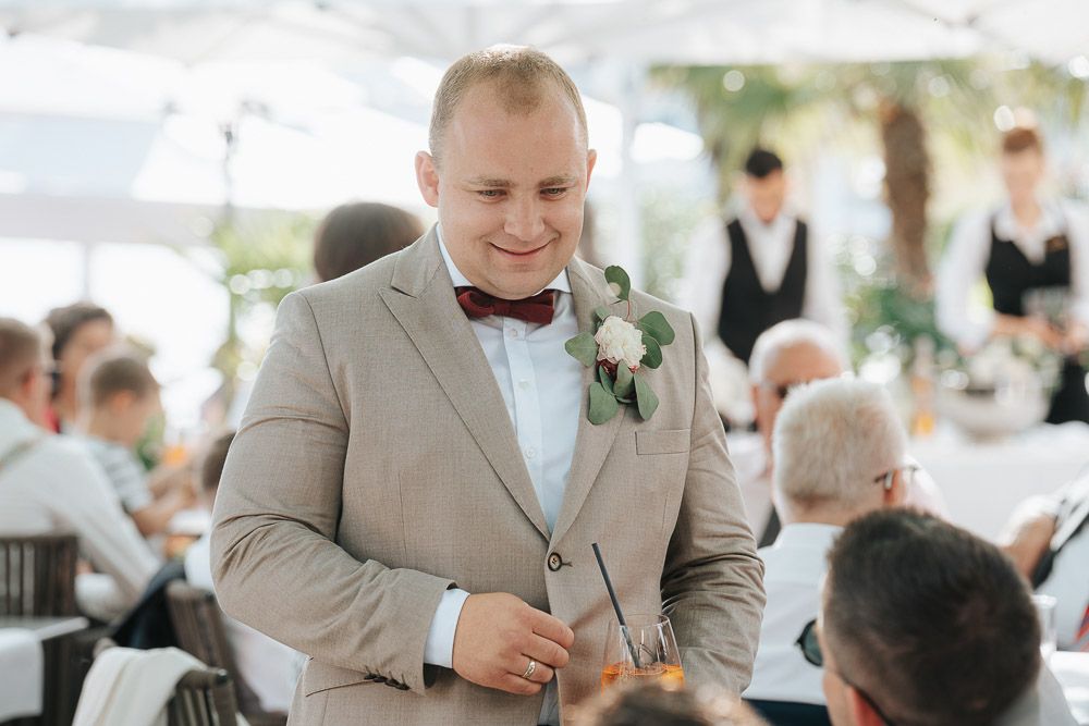 A person in a beige suit and bow tie smiles during an outdoor wedding reception under a tent.