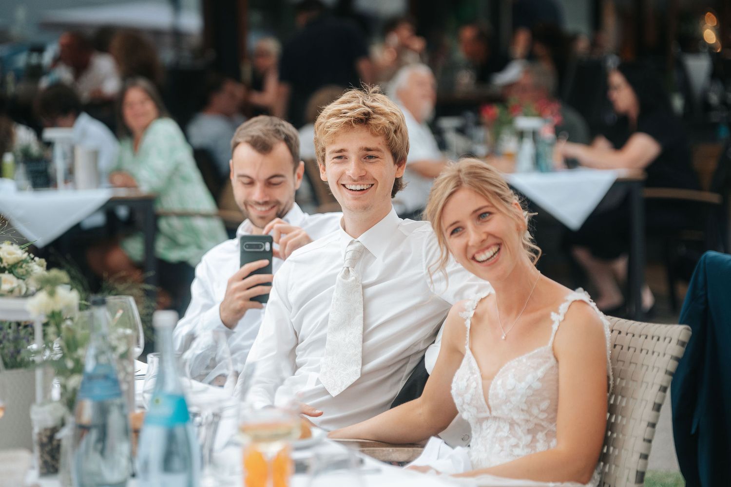 Smiling bride in a white lace dress sits at a reception table with two cheerful men in white shirts, one holding a phone.