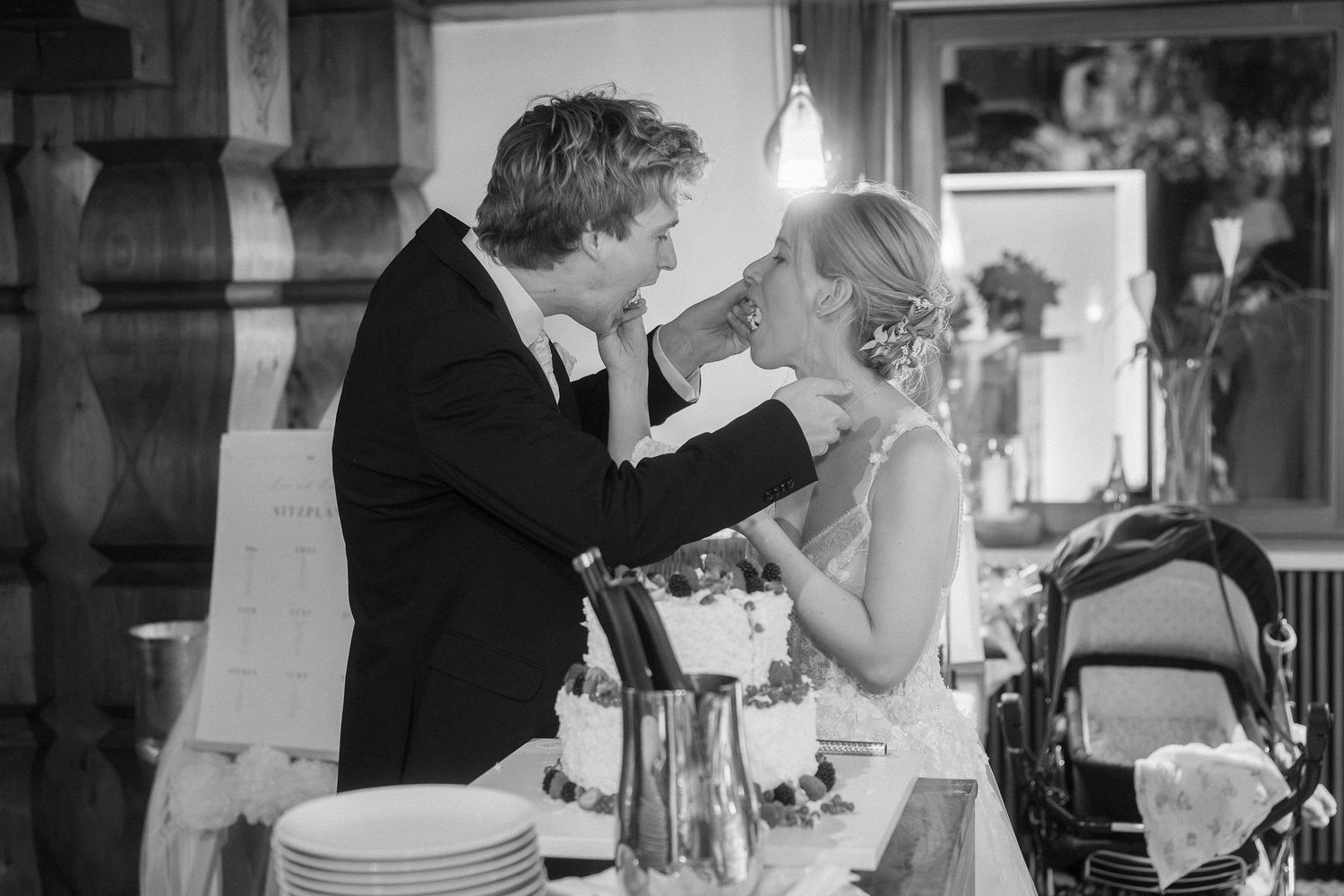 Black and white photo of a bride and groom playfully feeding each other wedding cake at their reception, with a stack of plates nearby.