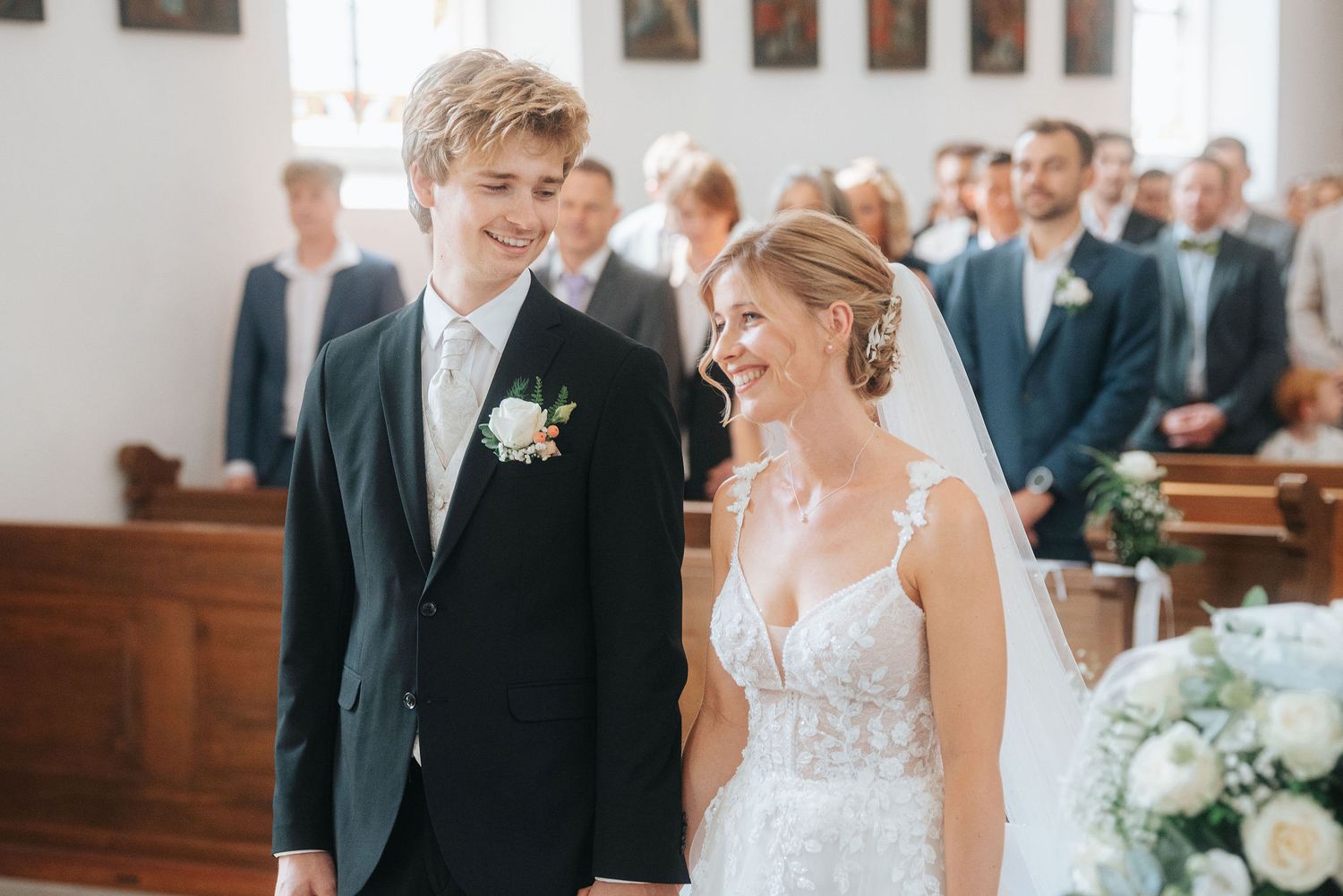 Smiling bride in lace gown and groom in dark suit walking down church aisle on their wedding day.