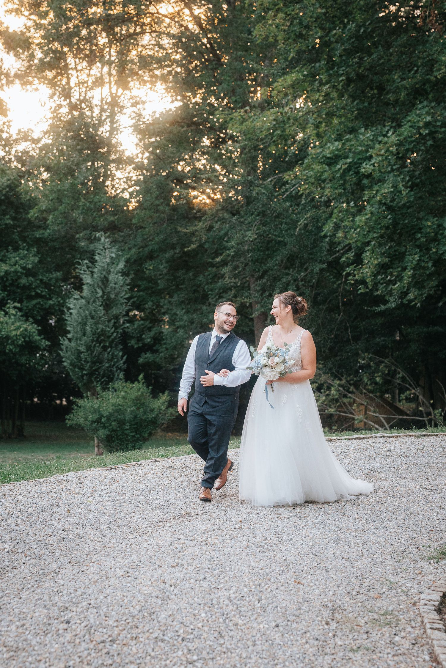Smiling bride in a white gown holding a bouquet walks beside her groom in a vest along a gravel path at sunset surrounded by trees.