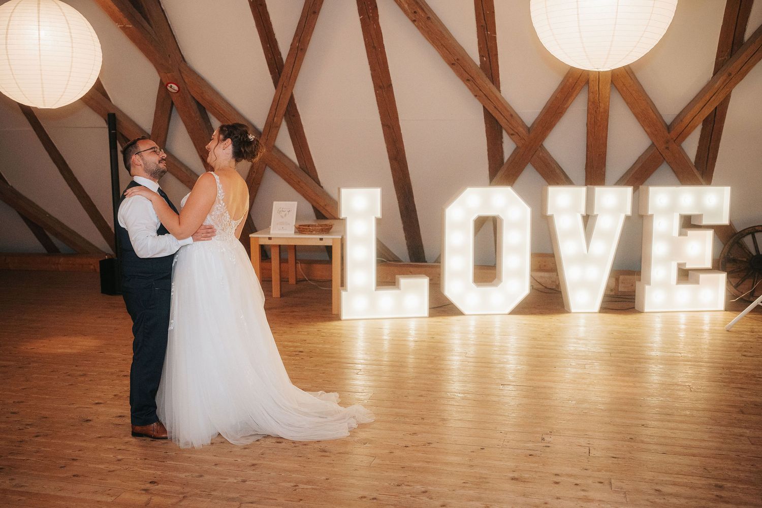 Bride and groom share an intimate dance beneath rustic wooden beams, with glowing marquee LOVE letters and warm string lights behind them.