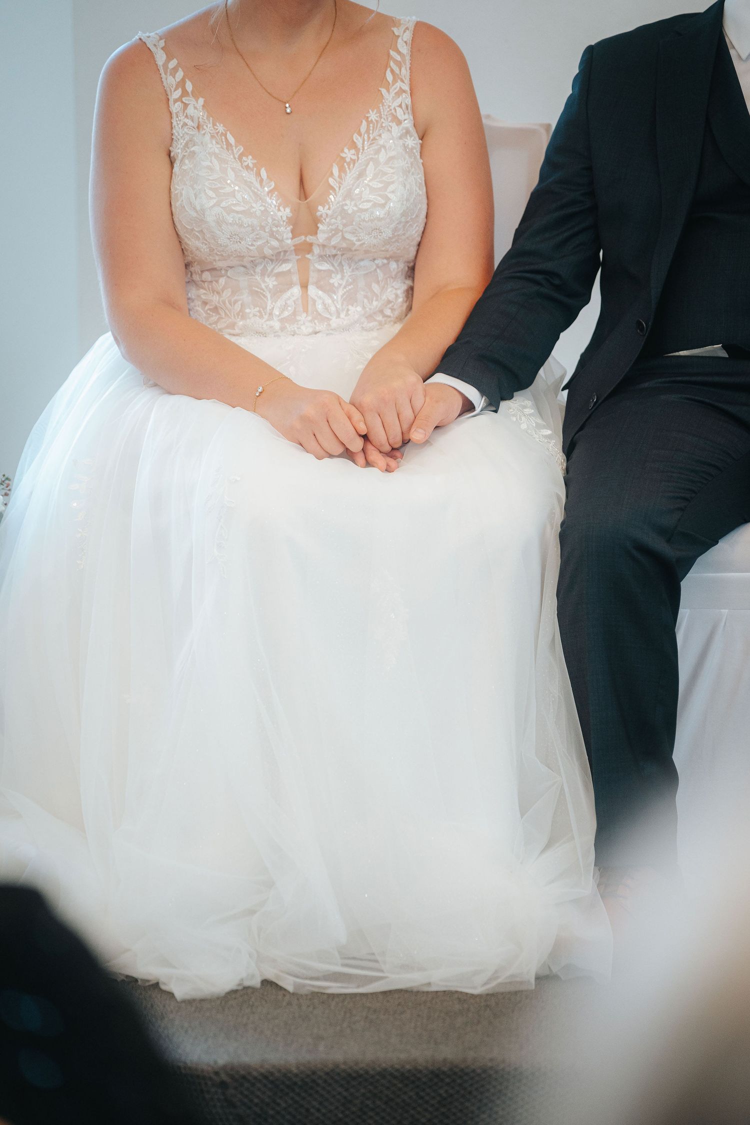 Bride in a white lace and tulle wedding gown holding hands with a groom in a dark suit while seated during a wedding ceremony.